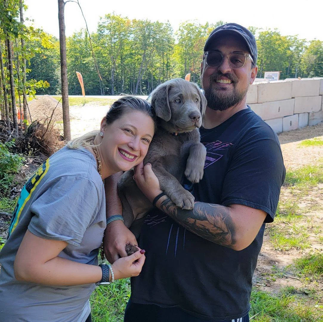A smiling woman and a man holding a gray Labrador puppy outdoors in a green, wooded area.