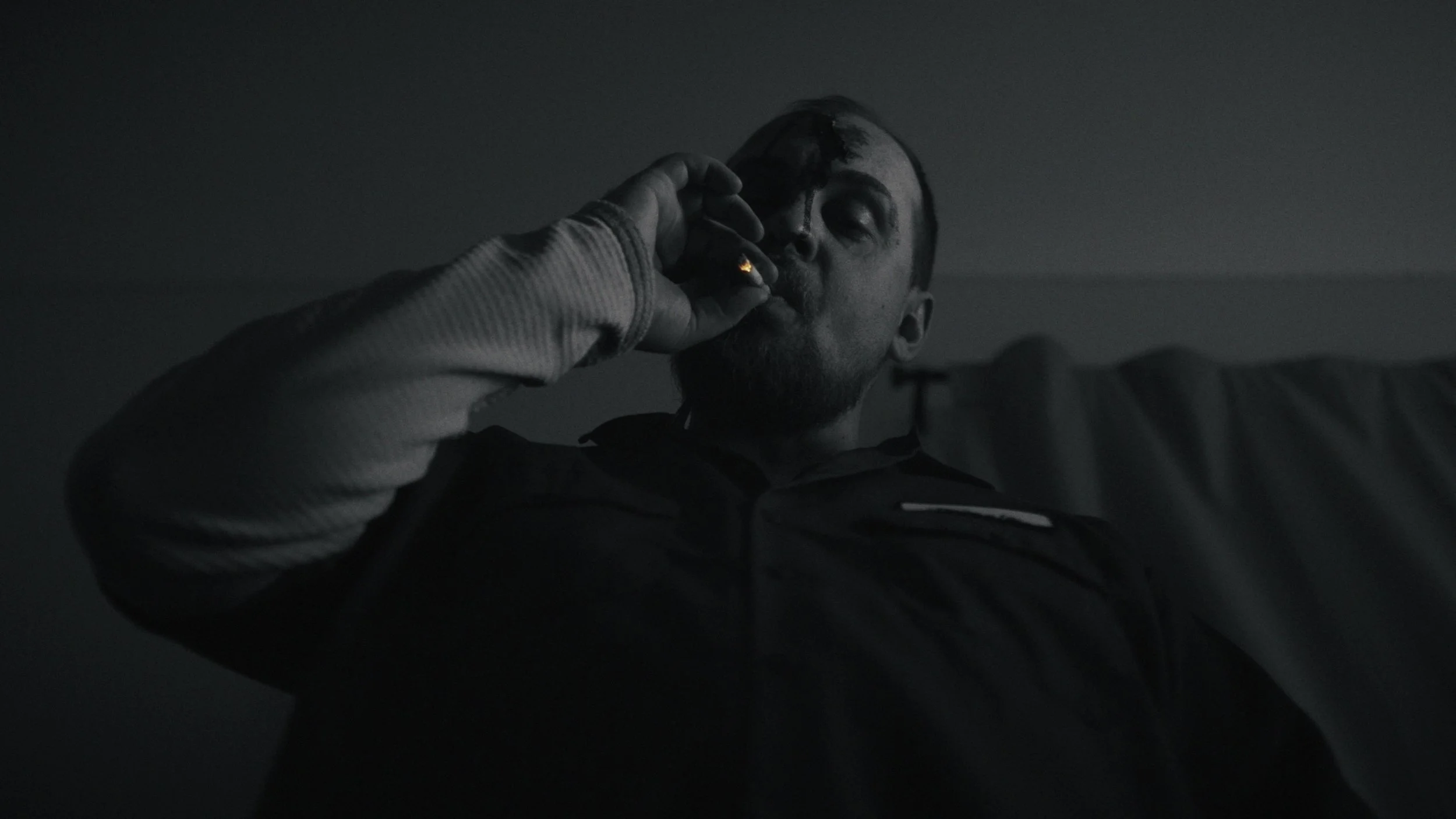 A man with a beard and short hair, wearing a jacket, is sitting in a dark room, smoking a cigarette.