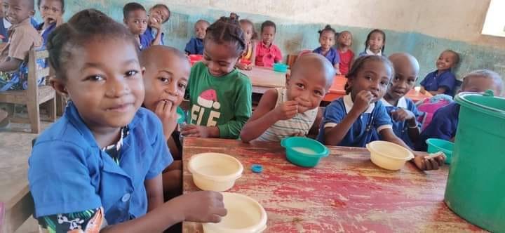 Children sitting at desks and smiling in a classroom, having a meal with bowls and cups.