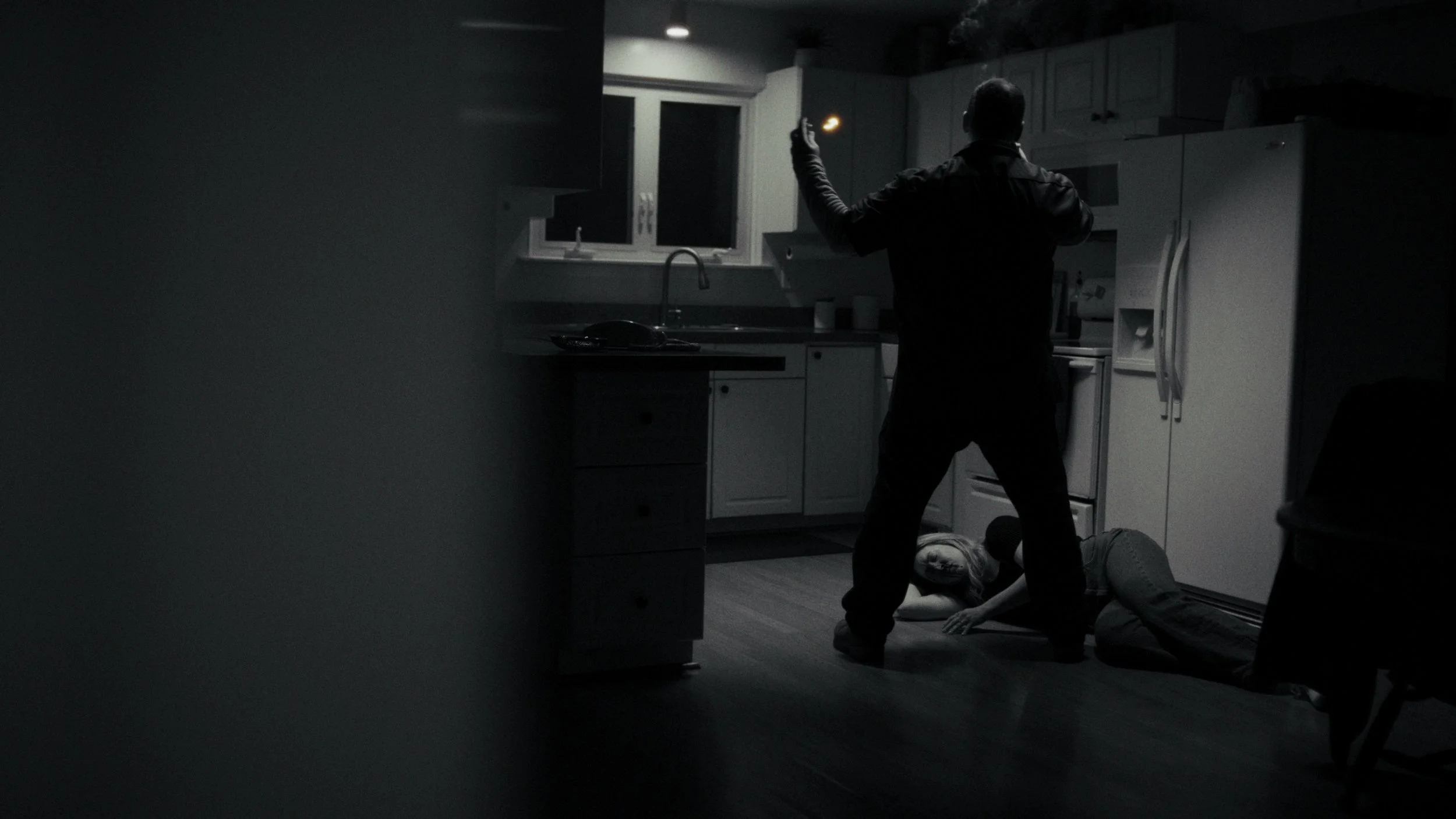 A man in dark clothing is standing in a dimly lit kitchen, holding a flashlight, with two people lying on the floor near his feet, one on her stomach and the other on her side, in a tense situation.