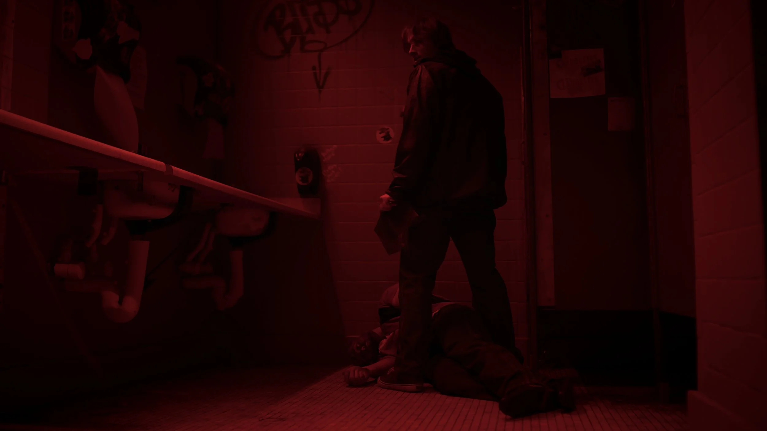 A person standing over another person lying on the floor in a dimly lit room with red lighting, graffiti on the wall, and sinks visible.