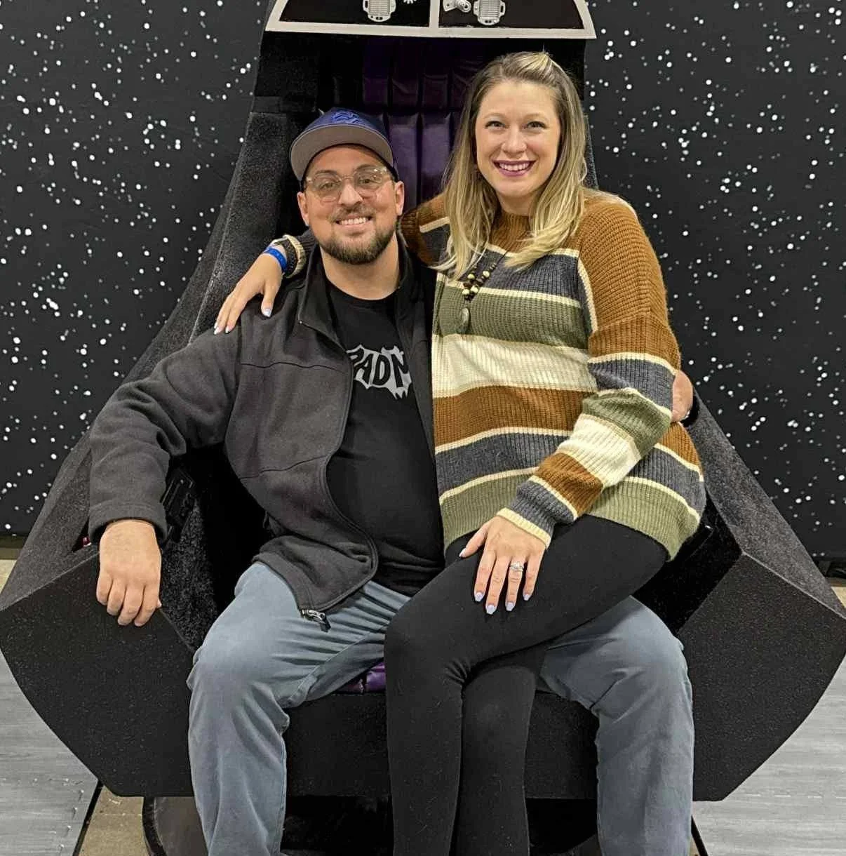 A man and woman sitting together on a black moon-shaped structure, smiling at the camera, with a starry space-themed backdrop behind them.