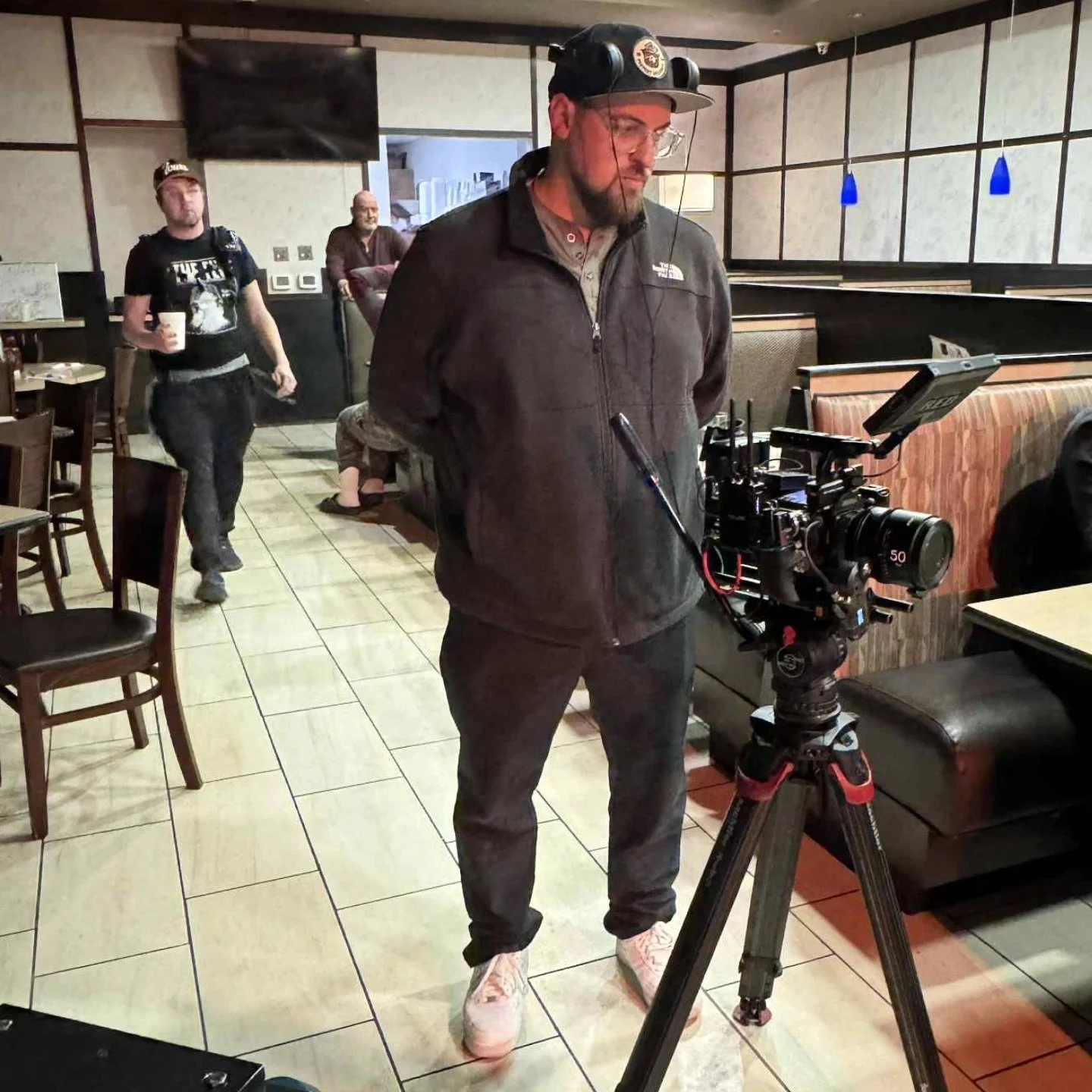 A man with glasses and a beard operating a professional video camera on a tripod inside a restaurant, with two other people in the background.