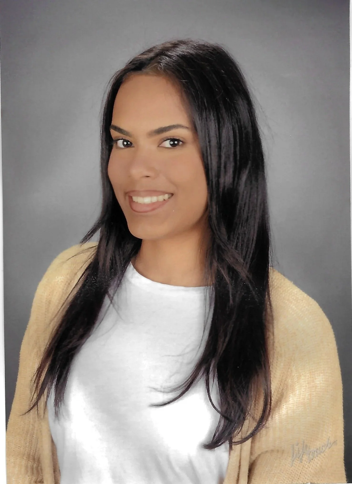A woman with long black hair, smiling, wearing a white shirt and a beige cardigan, standing against a plain gray background.