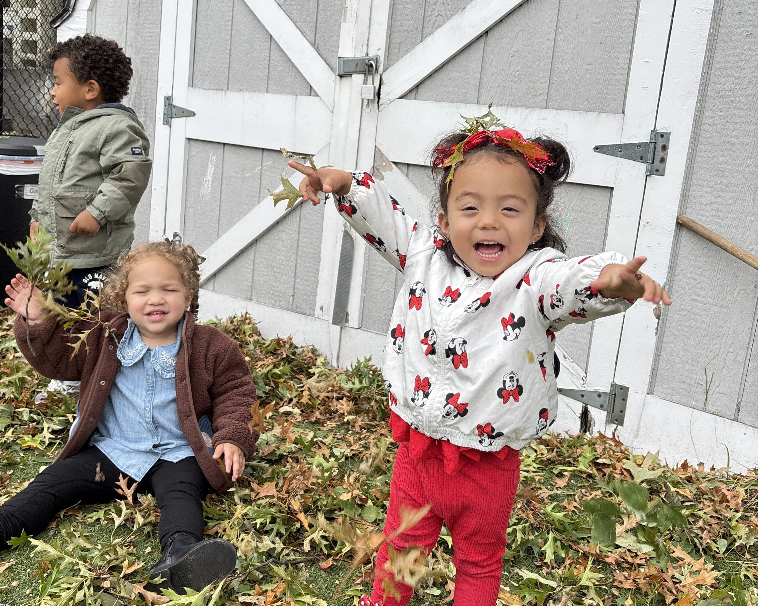Three children playing outside among fall leaves in front of a wooden shed. One girl in the foreground is smiling, wearing a Minnie Mouse jacket, red pants, and a holiday leaf headband. A girl in the background is sitting on the ground with a brown jacket and blue shirt, and a boy is standing in the background wearing gray with long sleeves.