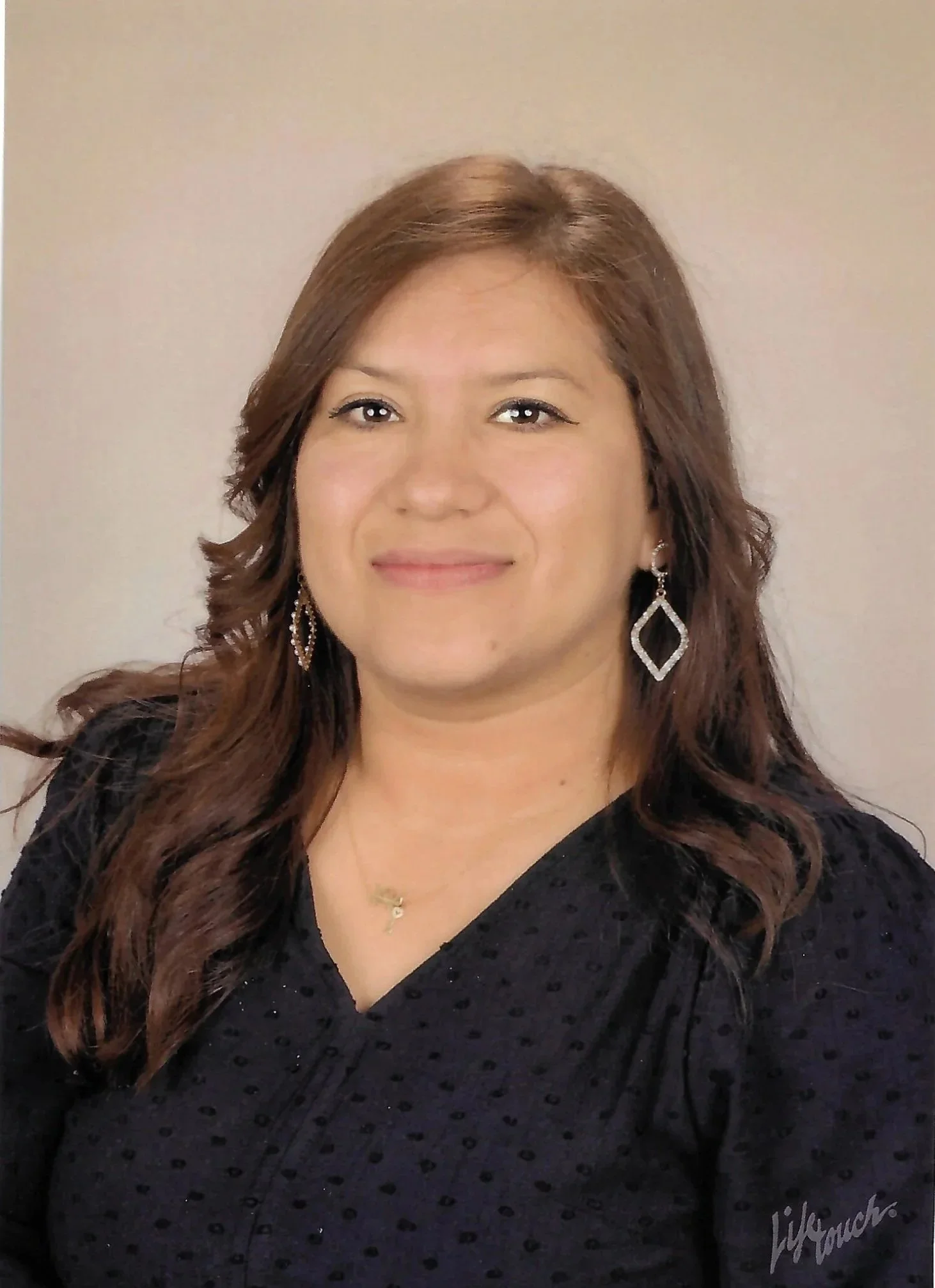 A woman with long brown hair, wearing a black textured top, hoop and diamond-shaped earrings, and a necklace, smiling at the camera against a plain beige background.