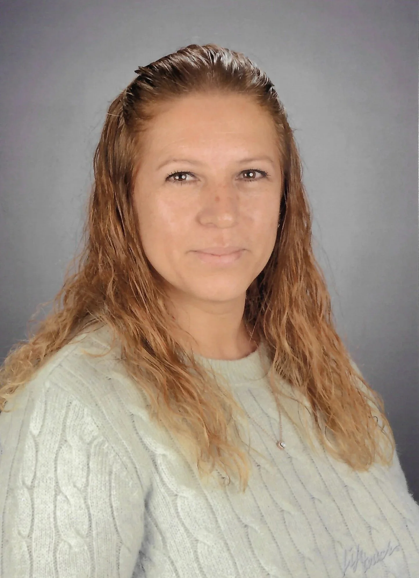 Woman with long, wavy red hair wearing a cream-colored sweater, looking at the camera with a neutral expression, against a gray background.