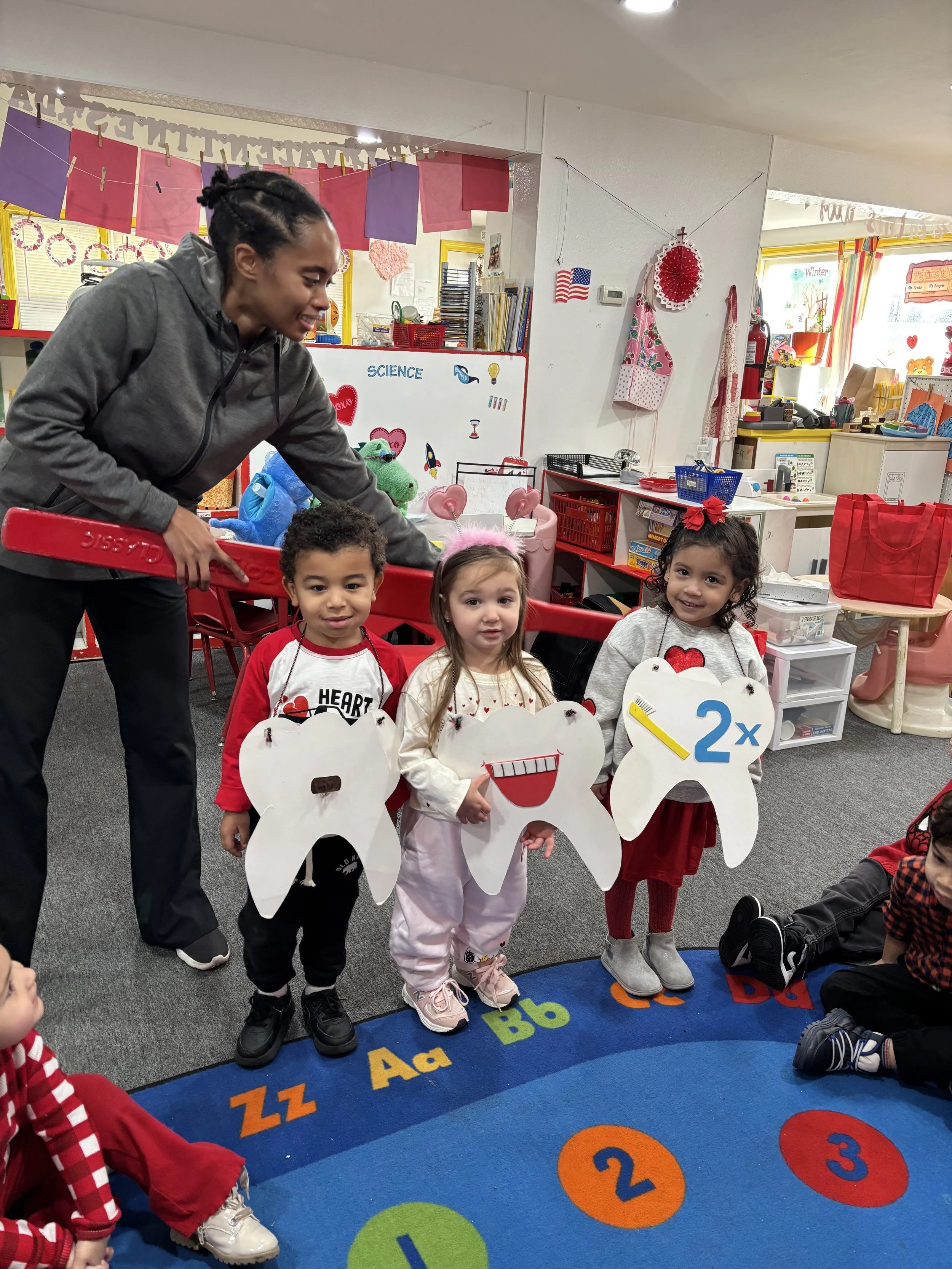 A teacher and three young children holding large paper teeth with dental-themed decorations, in a colorful preschool classroom.
