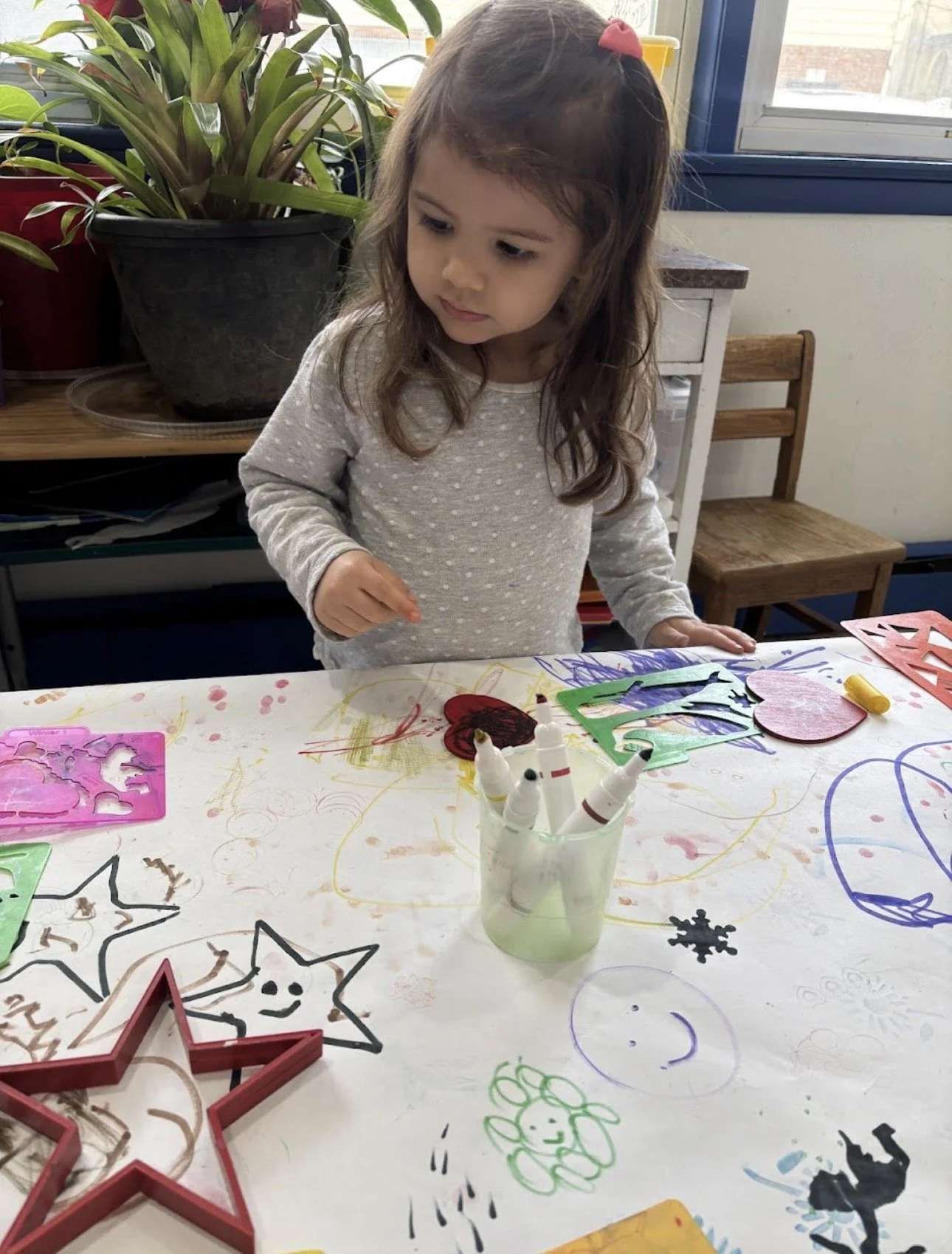 A young girl with brown hair, wearing a gray polka-dotted shirt, is standing at a table decorated with drawings and art supplies. She is looking down at the table, which has colorful papers, markers, stencils, and drawings of stars, hearts, and smiley faces.