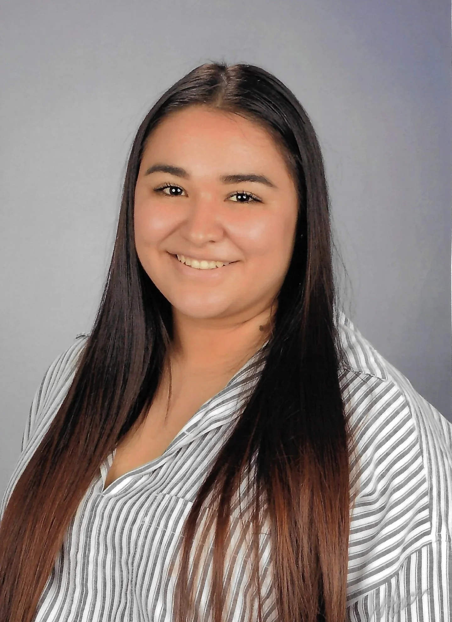 A woman with long black and brown hair, smiling at the camera, wearing a striped shirt, against a neutral background.