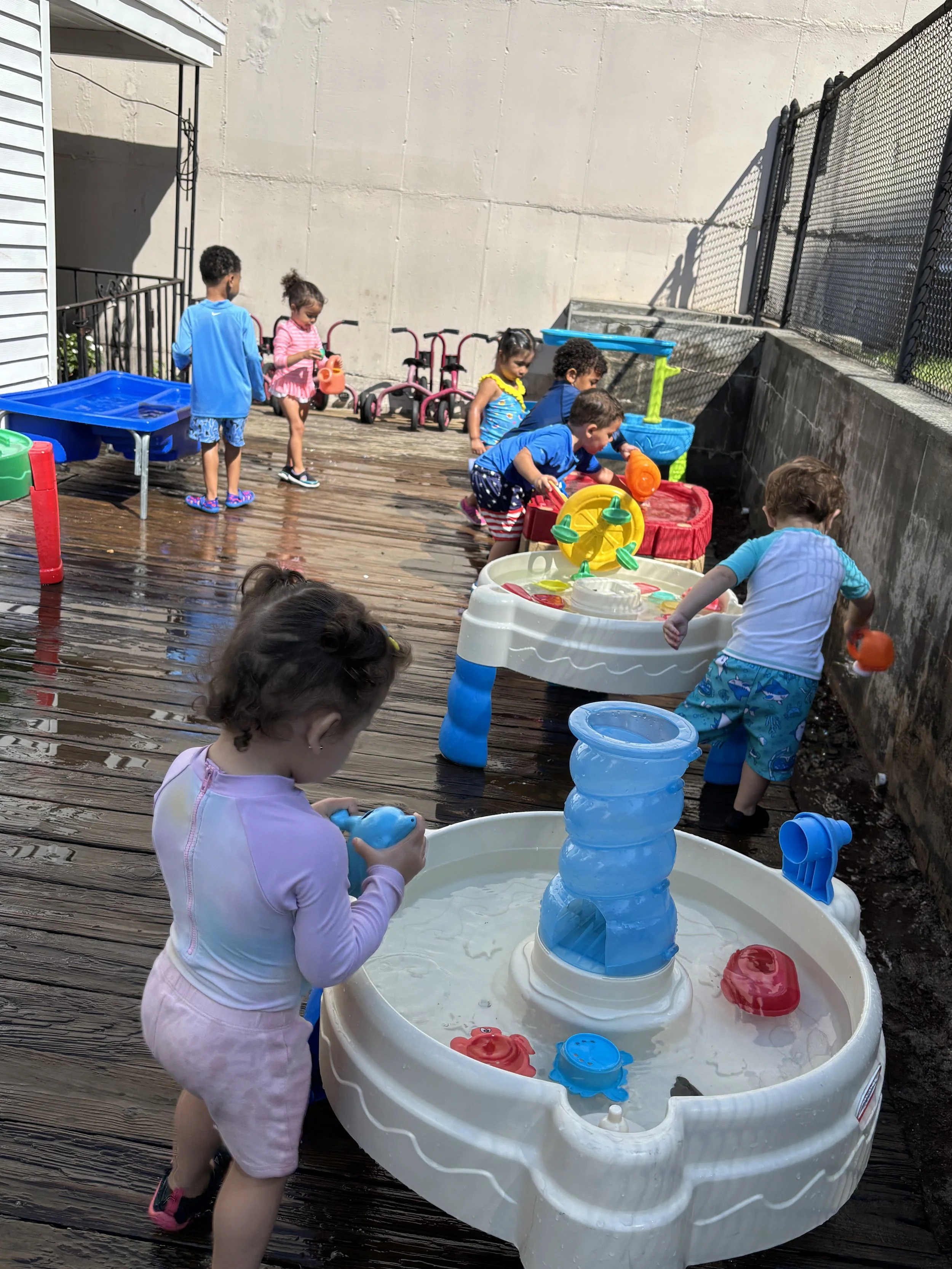 Children playing with water toys on a wooden deck outdoors, some are filling and playing in water tables, with various colorful toys and small tricycles in the background.