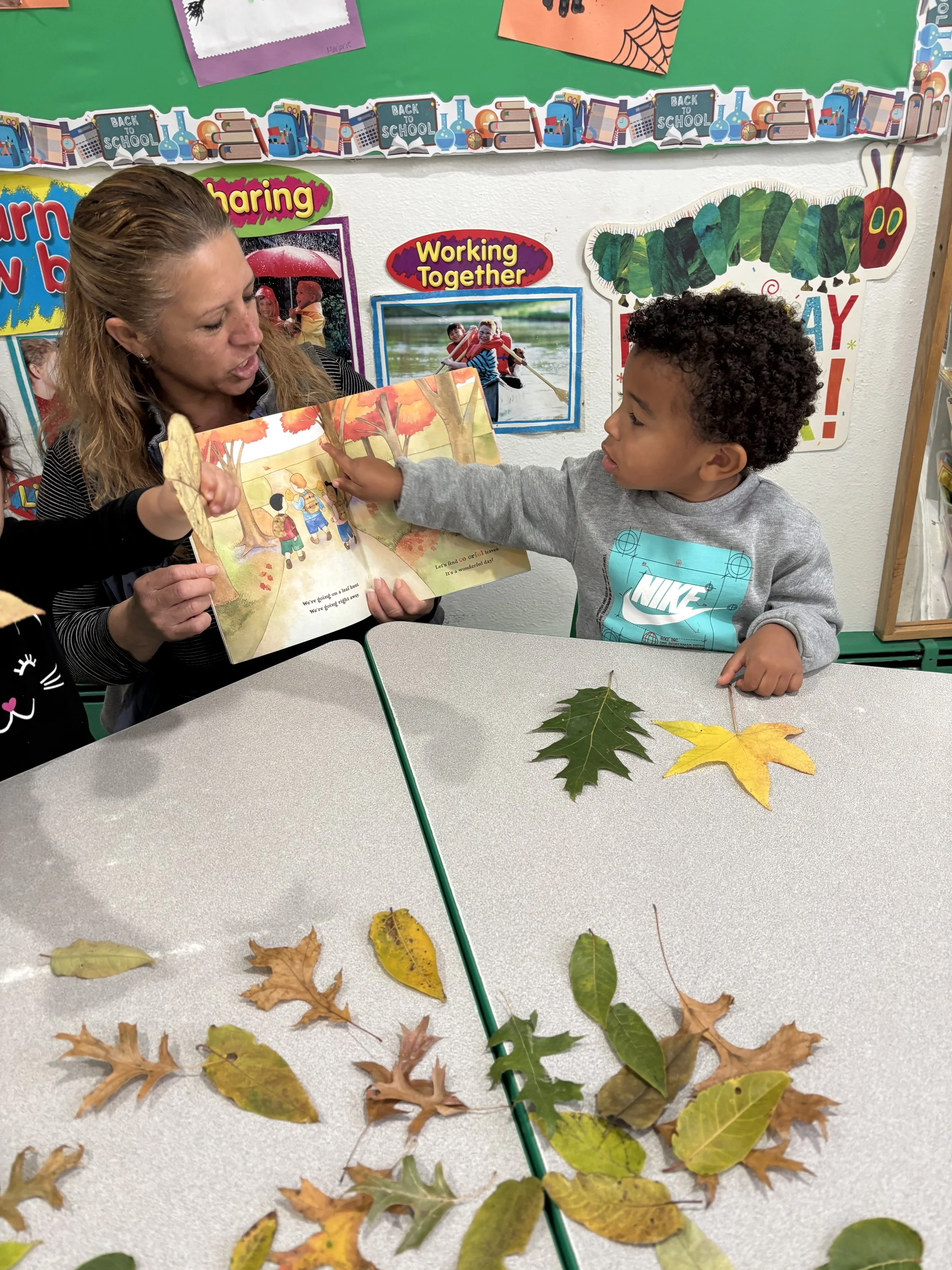 A teacher and a young boy sitting at a table with fallen autumn leaves, reading a children's book together. The background features a colorful classroom with educational posters and decorations.
