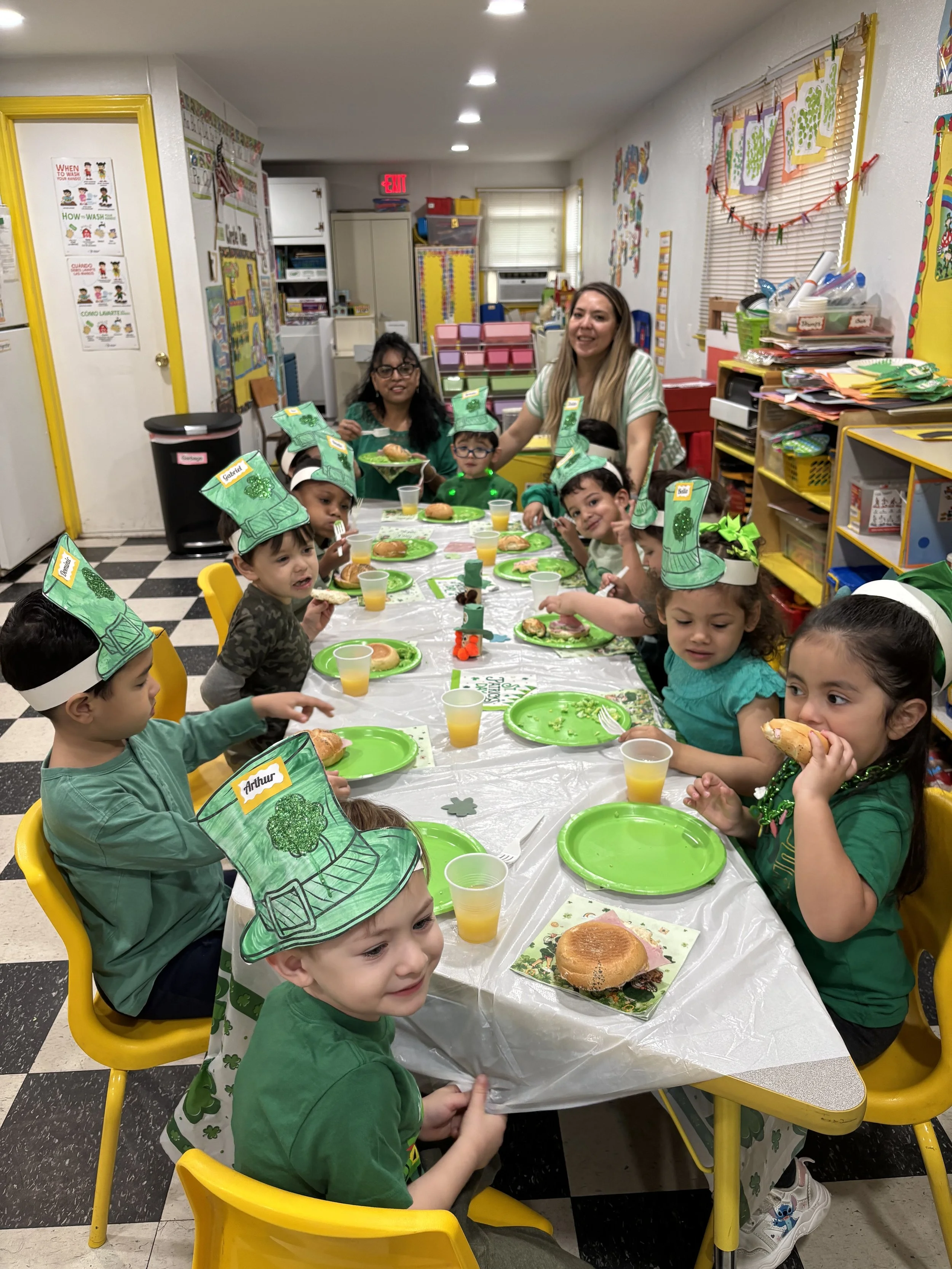 Children celebrating St. Patrick's Day at a daycare classroom party, wearing homemade leprechaun hats and eating cupcakes and sandwiches at a decorated table.
