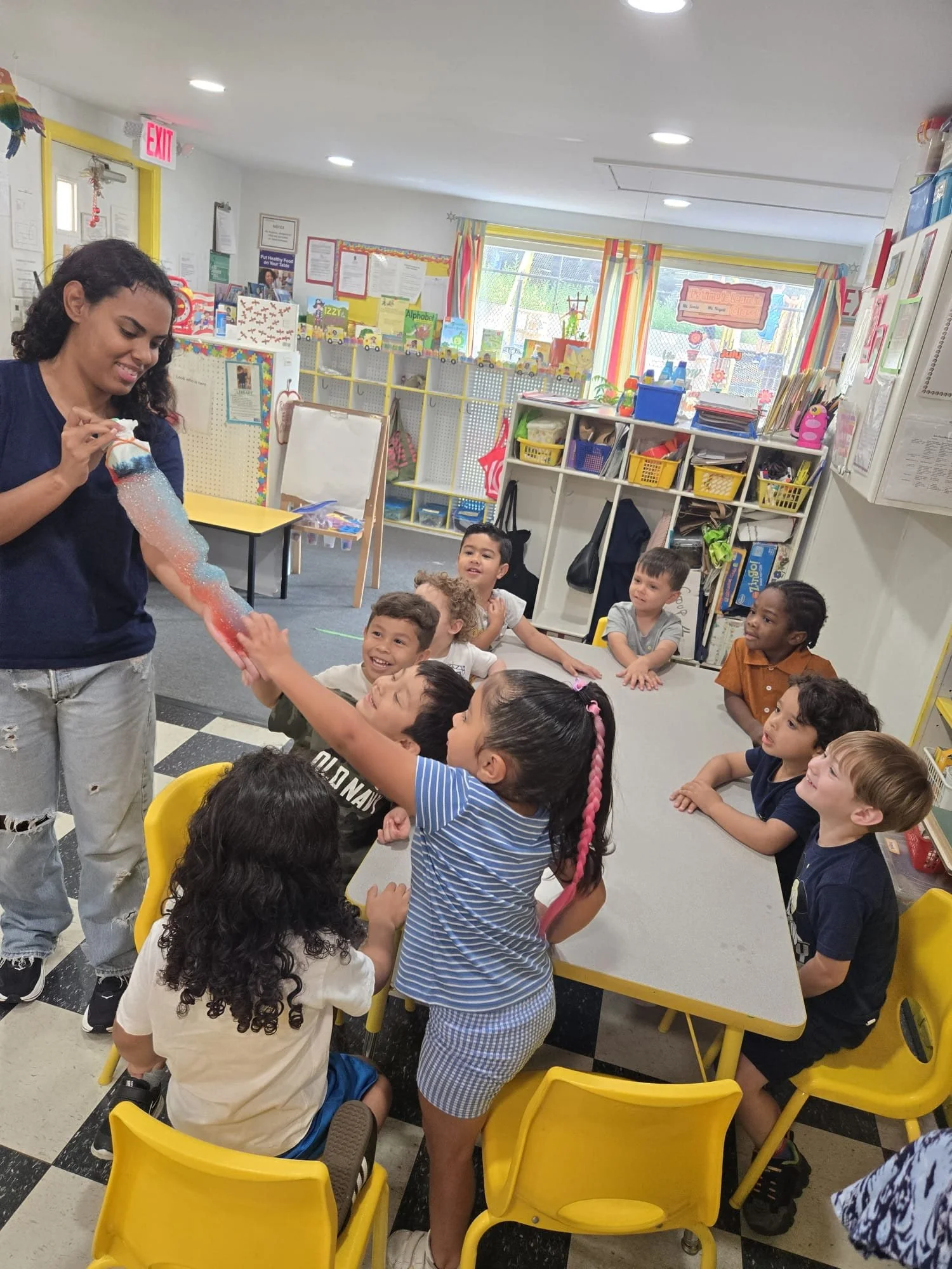A preschool teacher showing a colorful cotton candy to a group of children sitting at a yellow table in a classroom. The children are excited and reaching out for the cotton candy, with cheerful expressions.