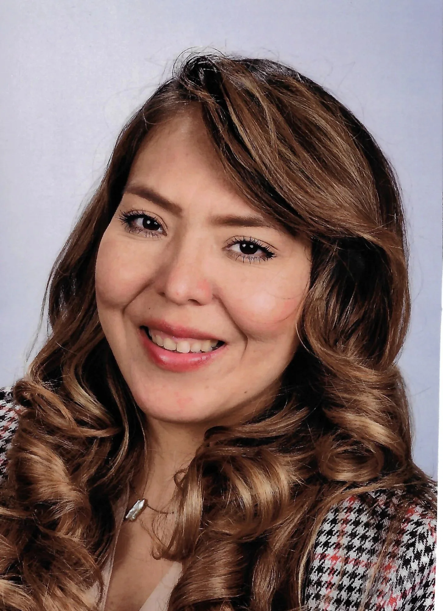 A woman with styled brown curly hair, wearing makeup, a necklace, and a checkered top, smiling for the camera against a plain background.