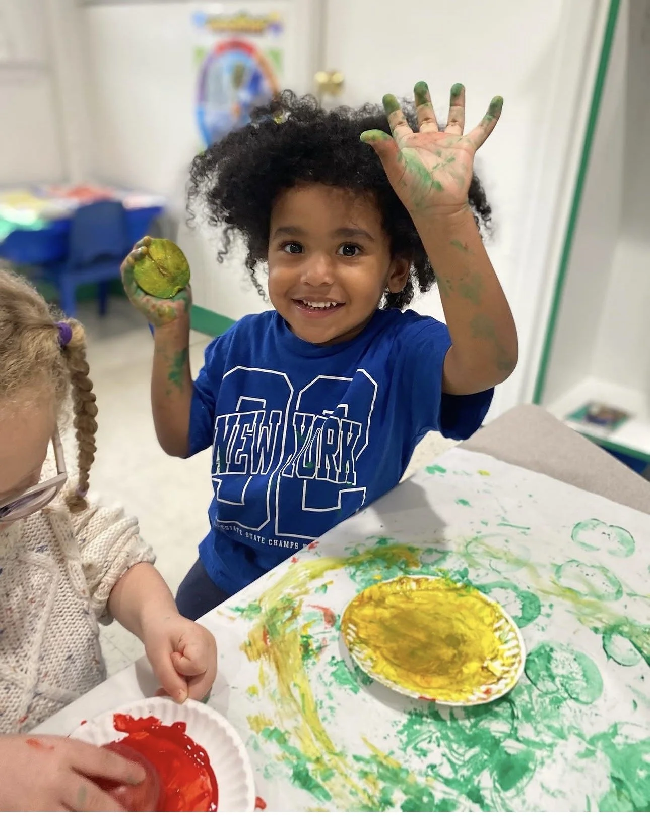 A smiling child with curly hair, wearing a blue 'New York' t-shirt, is holding a green painted object in one hand and raising the other hand, also painted green. The child is covered in green paint on the hands and arms. There are art supplies and a colorful paper plate with yellow paint on the table, and another child's arm painting with red paint is visible.