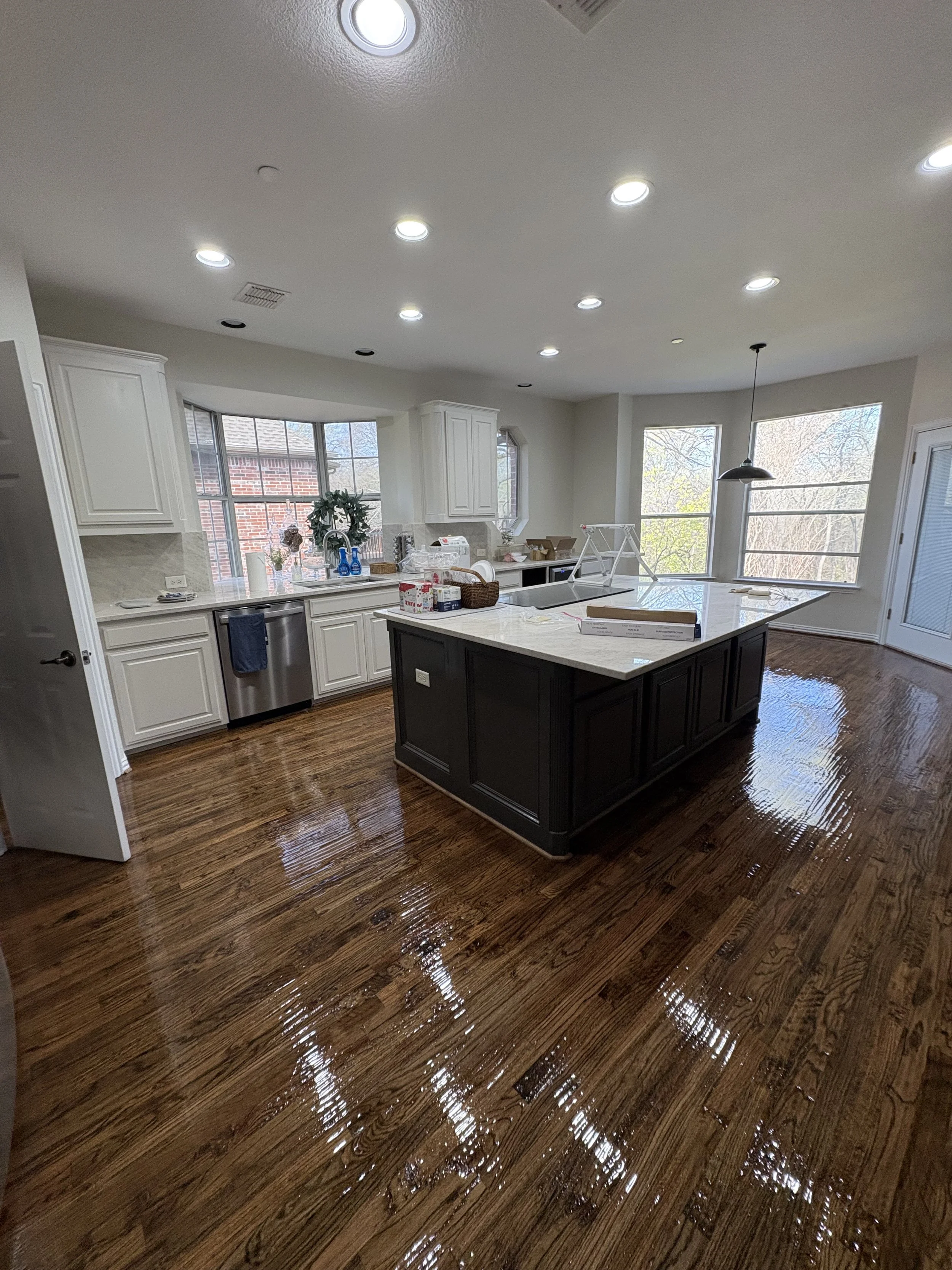 Bright modern kitchen with white cabinets, a dark central island, and shiny hardwood floors, sunlight coming through large windows.