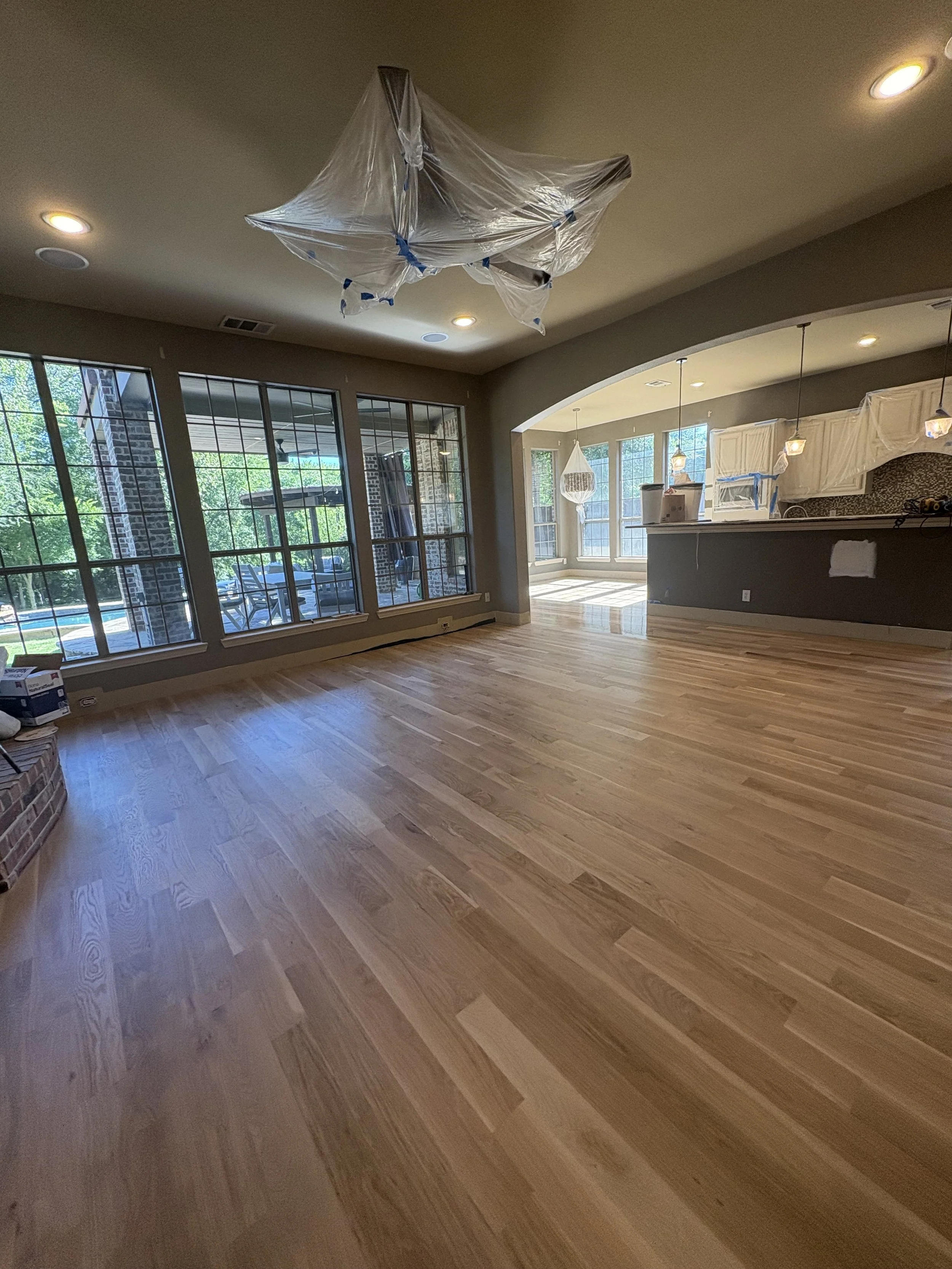 Empty living room with newly installed hardwood floors, large windows, and a chandelier wrapped in plastic hanging from the ceiling, indicating ongoing renovation.