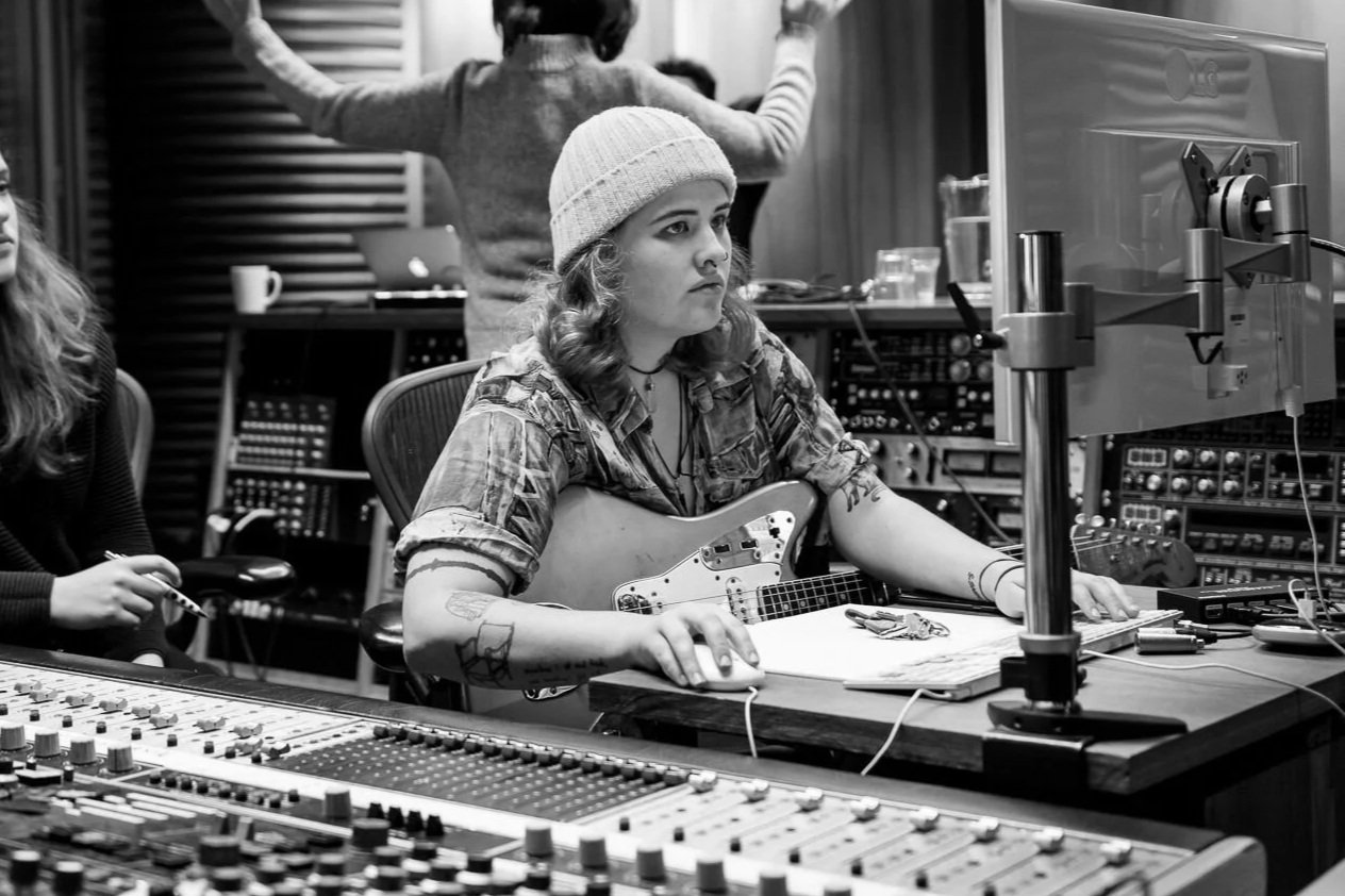 A woman with curly hair and a beanie wearing a patterned shirt, sitting at a recording studio with a guitar, computer, and audio equipment, focused on a monitor.
