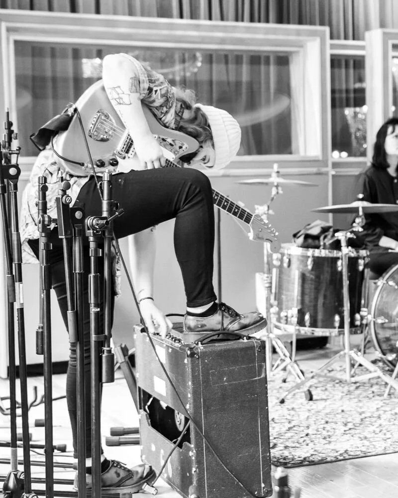 Black and white photo of a woman playing an electric guitar with her foot on an amplifier, in a music studio with drums and a woman in the background.