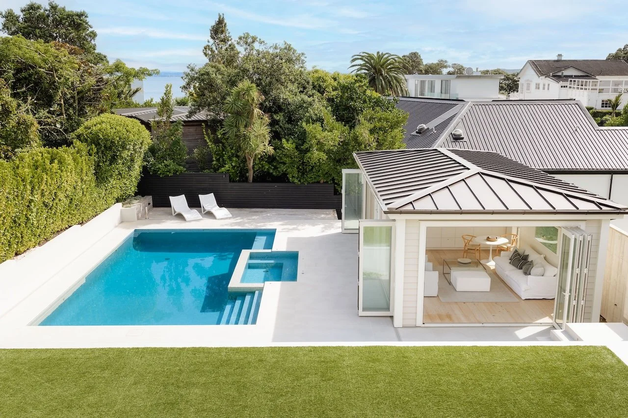 Looking from 20 Springcombe Road, St Heliers, Auckland, across landscaped green space to the heated swimming pool and pool house. Sunlight, greenery and seamless indoor-outdoor living create a private setting for relaxation, entertaining and everyday