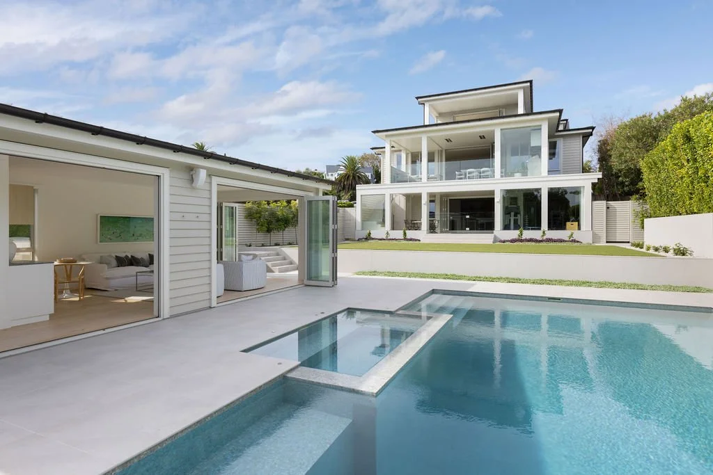 Looking across the swimming pool towards 20 Springcombe Road, St Heliers, Auckland, with the pool house opened fully to the outdoors beneath clear blue skies. Seamless connections between architecture, water and open-air living create a private envir