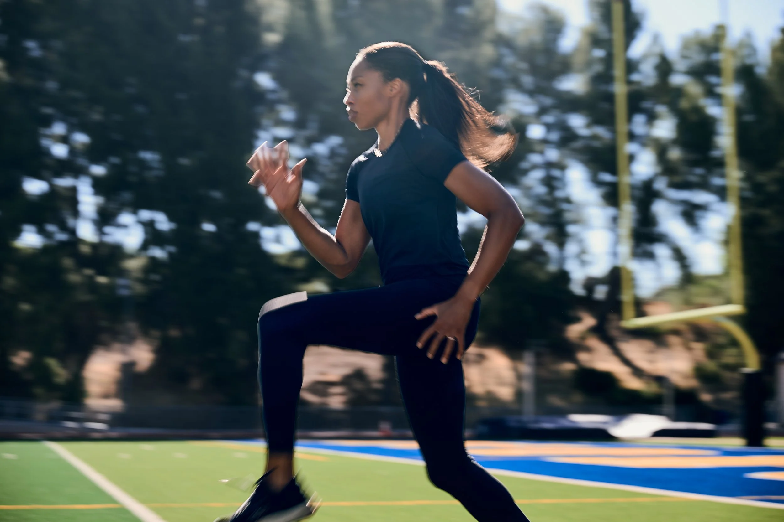 Allyson Felix running outdoors on a sports field in a side profile view, wearing a black athletic outfit and black athletic shoes, surrounded by trees and yellow goal posts.