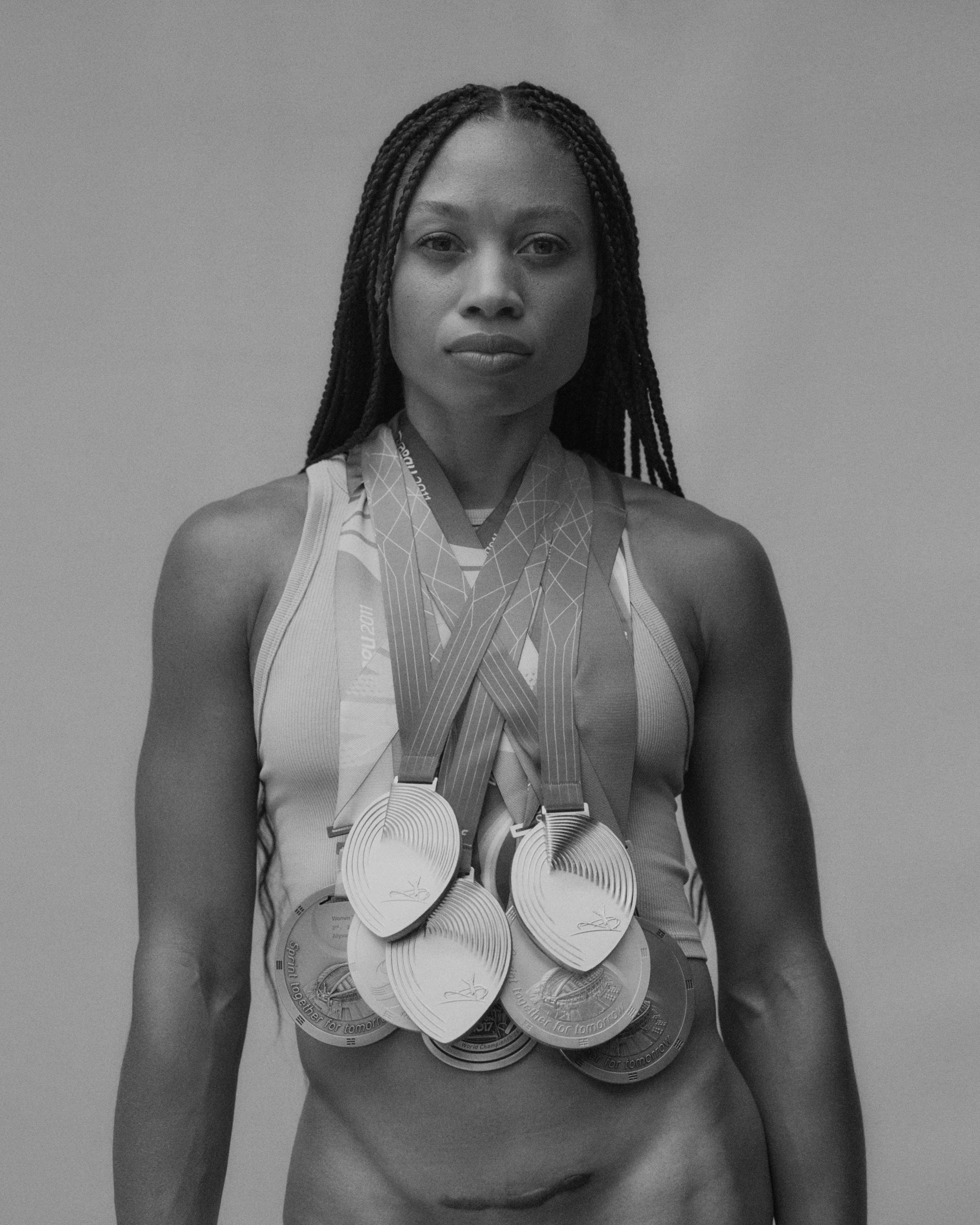 A female athlete with braided hair and a determined expression wearing a tank top and multiple medals around her neck, standing against a plain background.