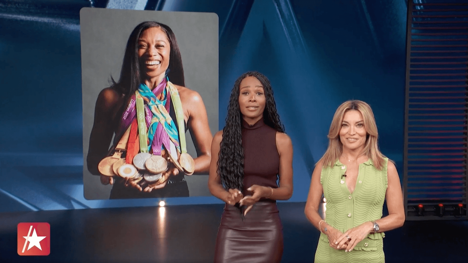 Two women are standing in a studio with a large screen behind them of holding numerous medals. The woman on the left has long dark braided hair and is wearing a sleeveless top and a leather skirt. The woman on the right has shoulder-length blonde hair and is wearing a green sleeveless vest with a watch and rings. The studio has dark blue walls and stage lighting.