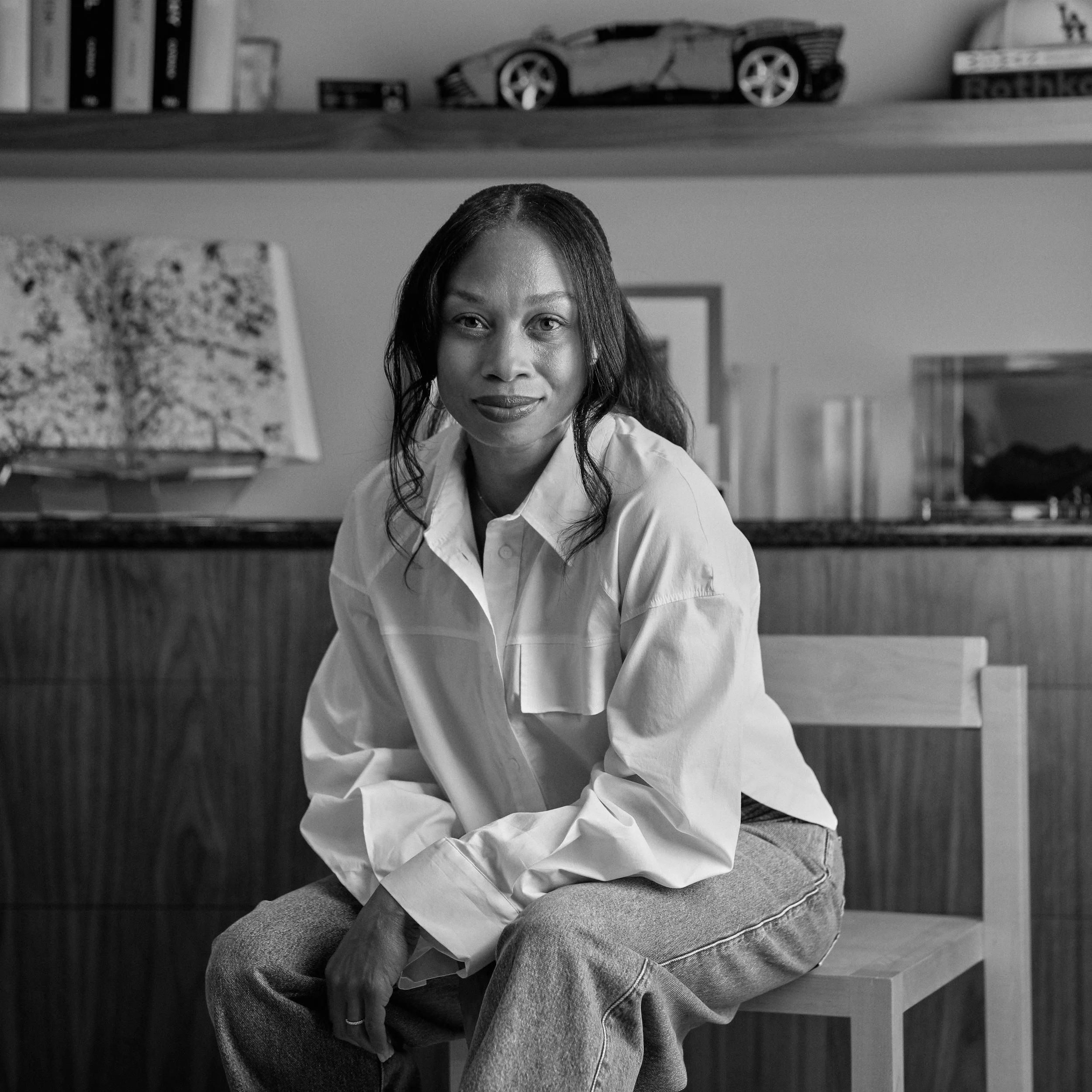 A woman with dark hair, wearing a white shirt and jeans, sitting on a chair with her leg crossed, in an indoor setting with shelves and a model car on the top shelf.