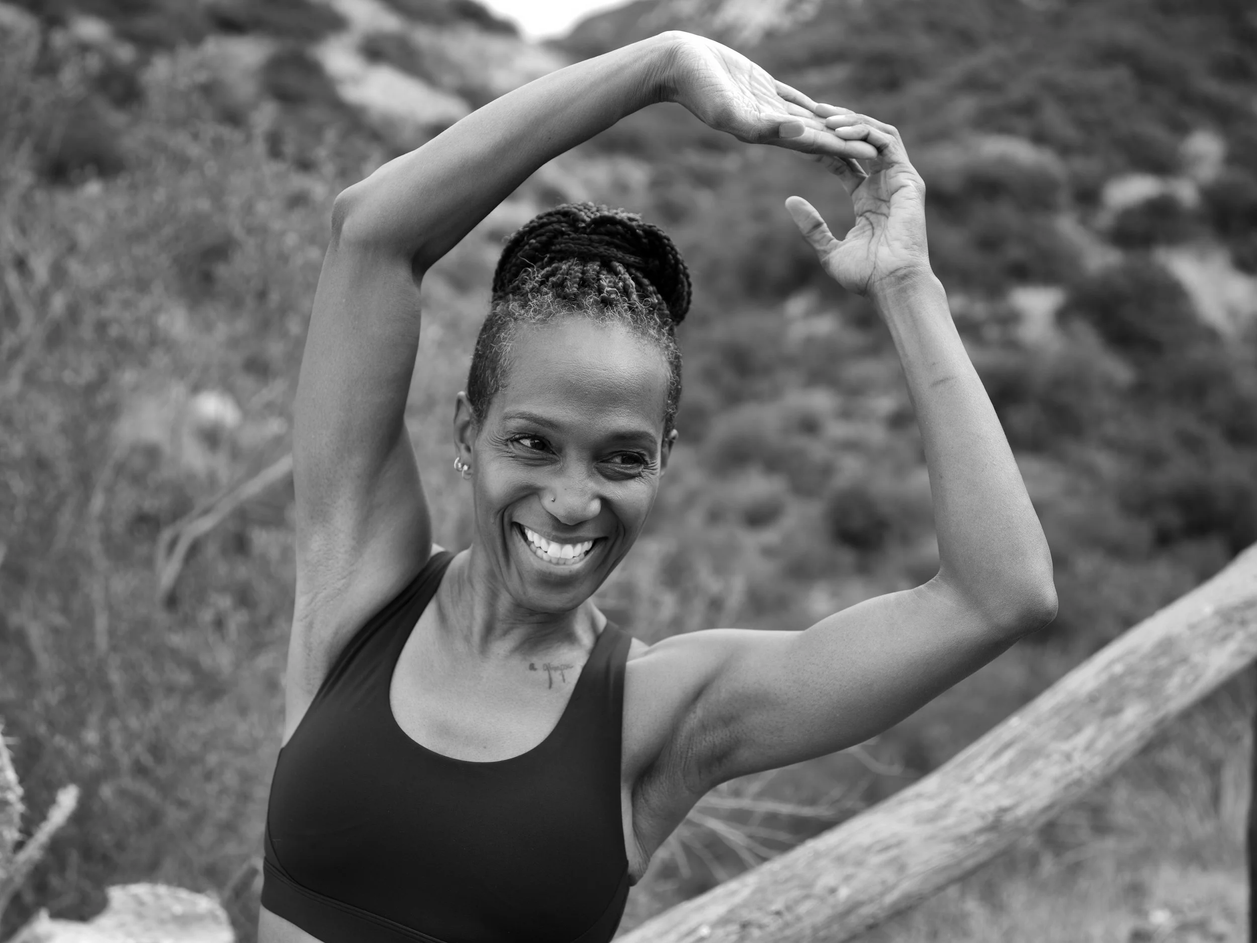 A smiling Black woman with braided hair stretching outside in a natural setting.