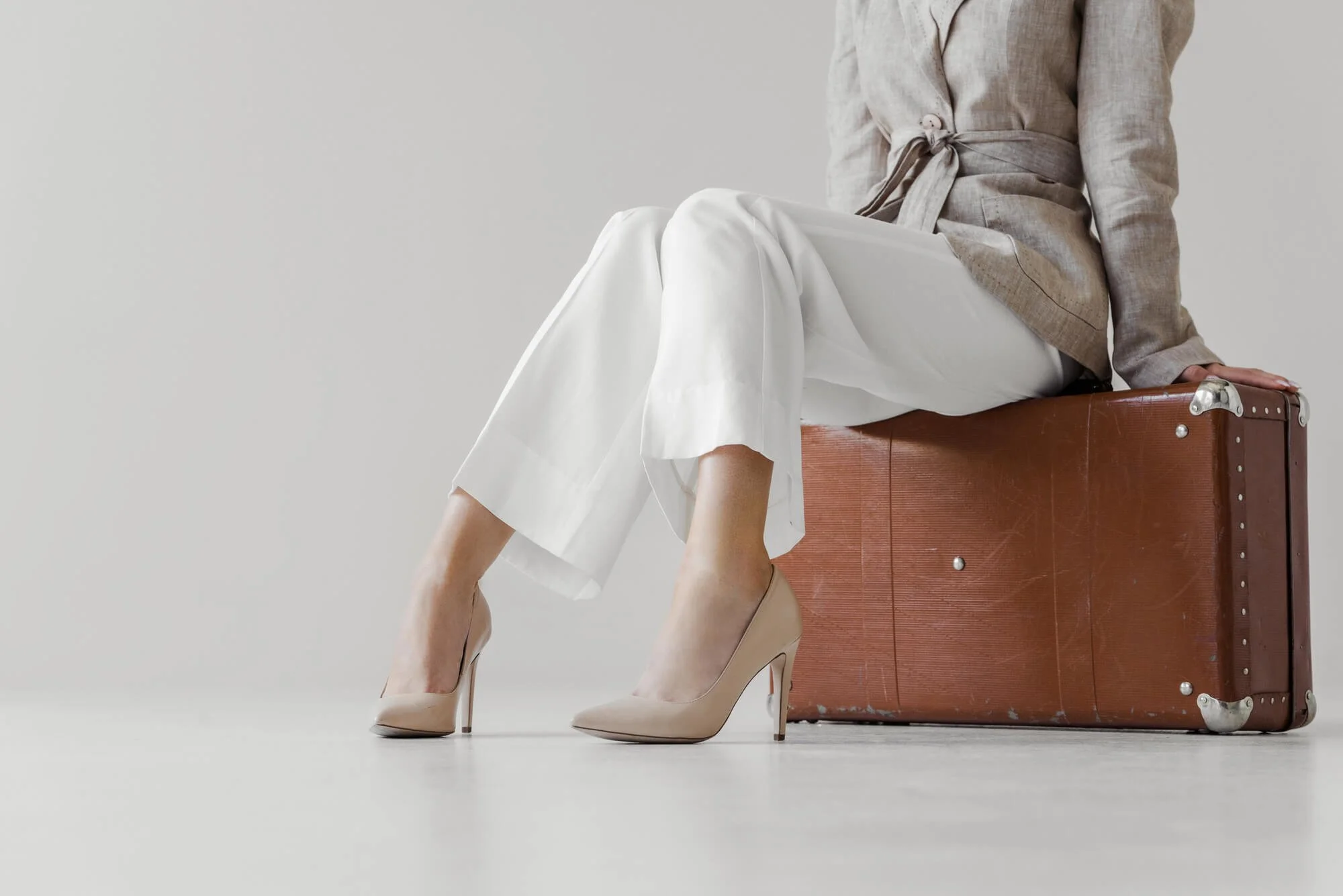 woman sitting on suitcase in heels, white pants and taupe blazer