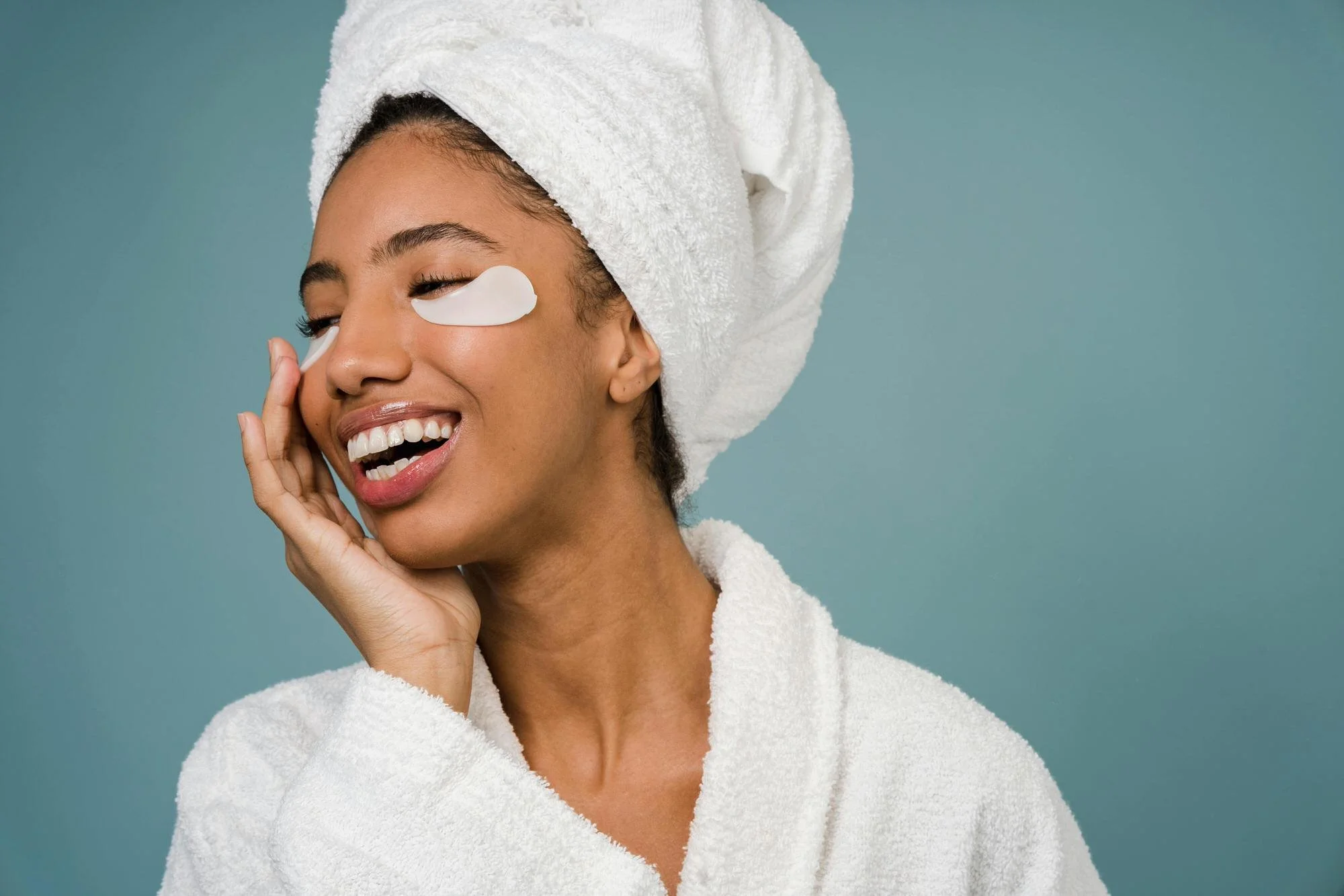 woman in bathrobe and head towel and eye patches smiling