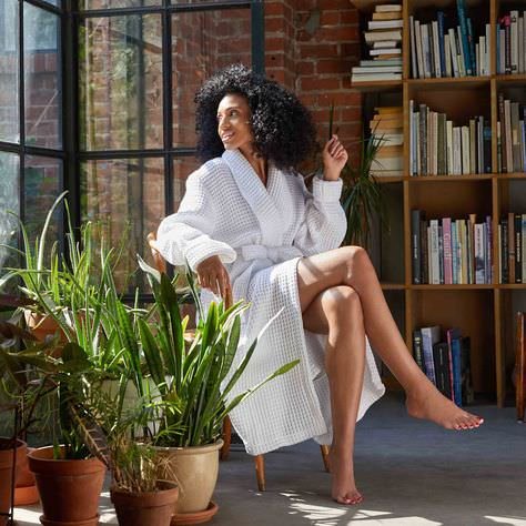 woman in bathrobe sitting legs crossed on chait in front of book case plants all around her