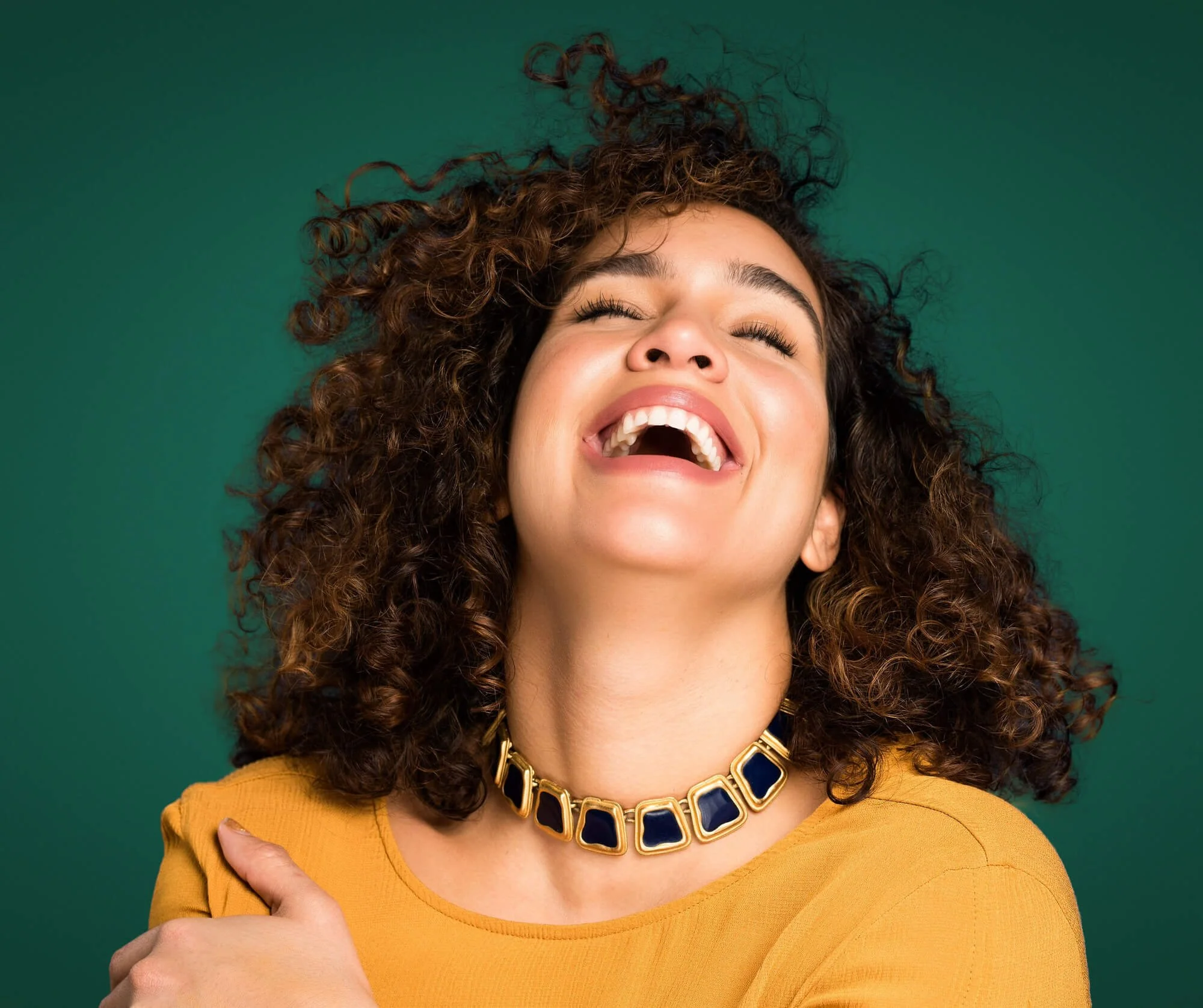 Dariana Casado - portrait laughing headback, big smile, eyes closed, green background yellow top, blue and gold necklace