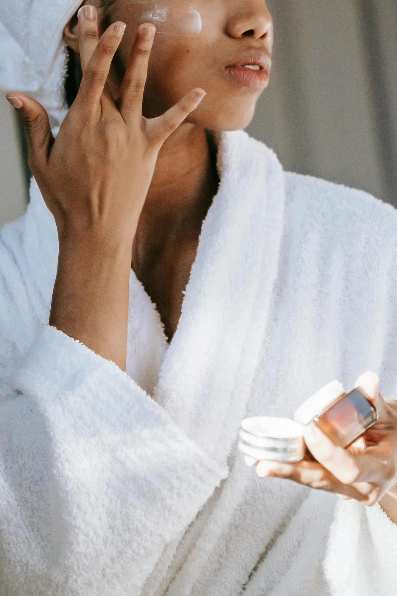 woman in white robe and white towel around her hair applying beauty cream to face