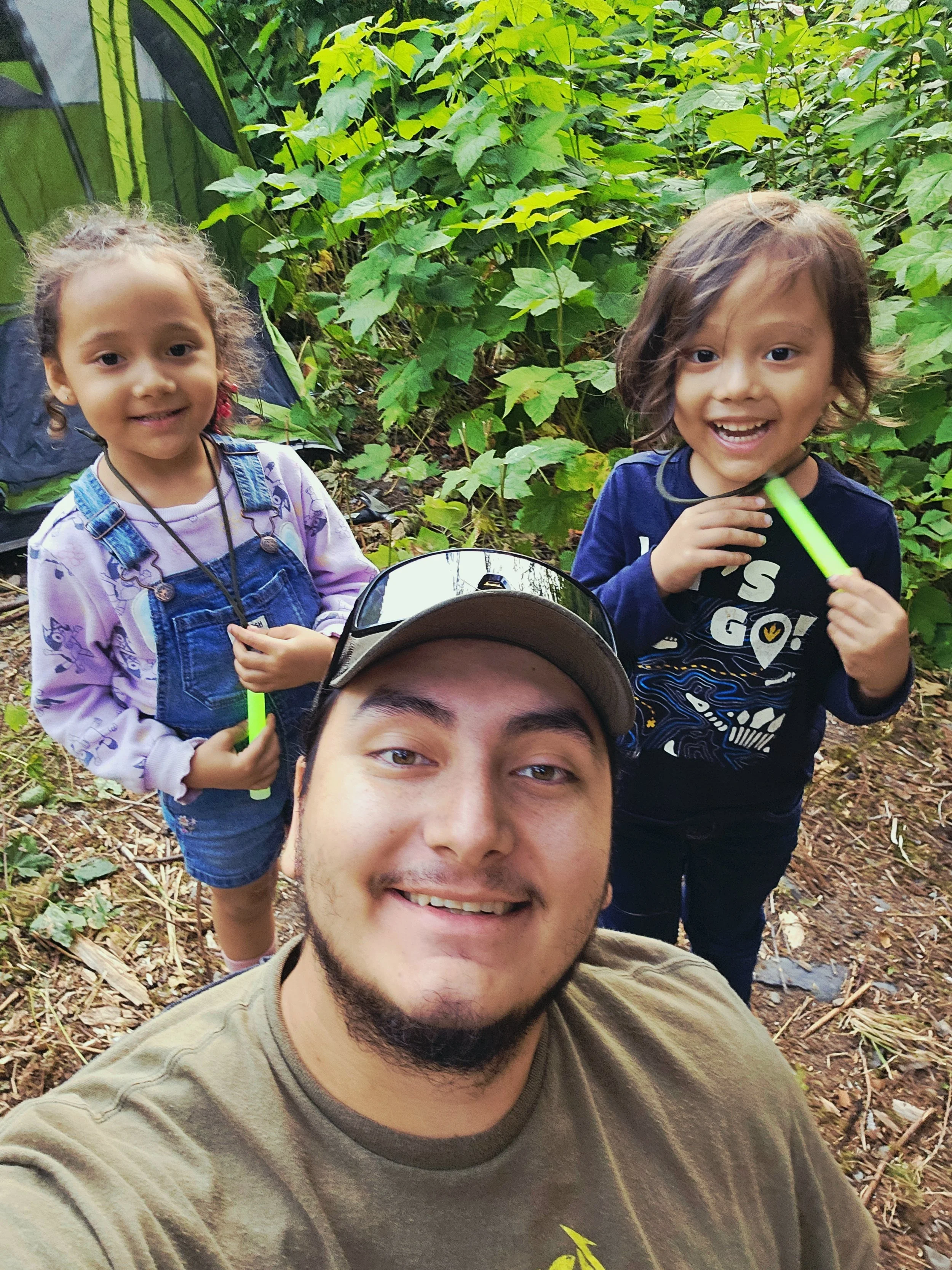 A man taking a selfie with two young children in a lush, green outdoor setting. The kids are smiling, and the man is wearing a baseball cap and sunglasses on his head.