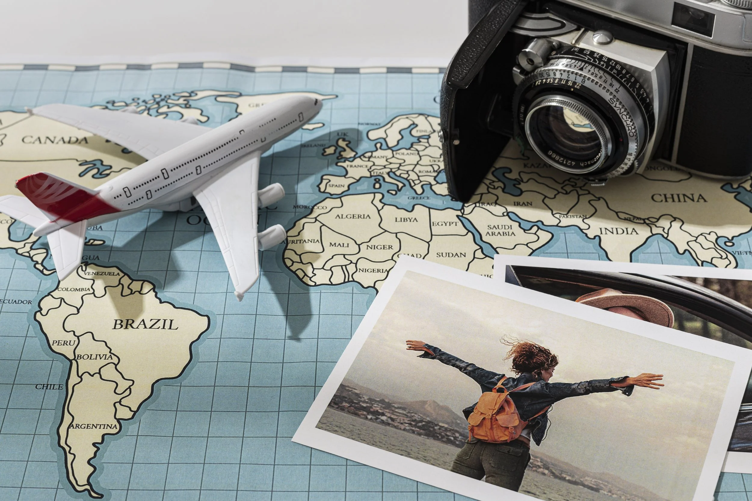 A world map with a toy airplane, a vintage camera, and a travel photograph of a woman with outstretched arms at the beach.