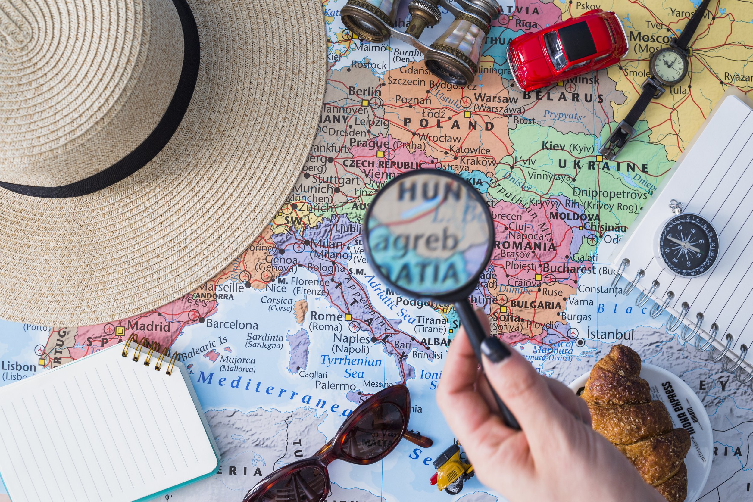 A world map displaying Europe and parts of Asia with items on top including a straw hat, a spiral notepad, sunglasses, a magnifying glass focusing on Greece, a pair of binoculars, a toy red car, a pocket compass, a small toy explosion, a clipboard with paper, and a bowl of croissants.