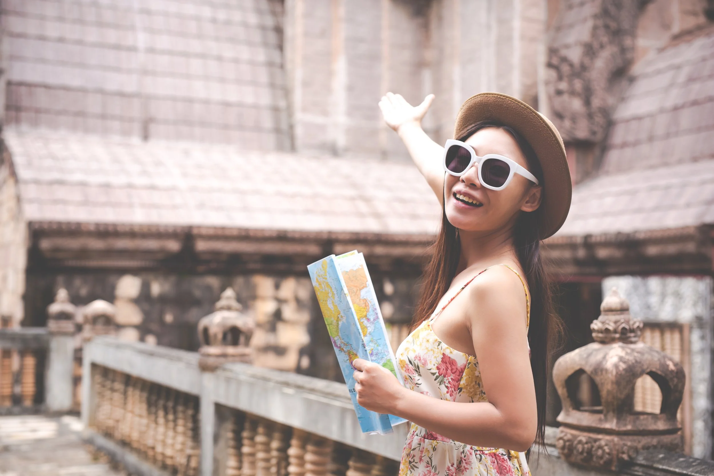 A woman with long brown hair wearing a hat and sunglasses, holding a map, smiling, and gesturing with one arm, in front of an ancient brick building.