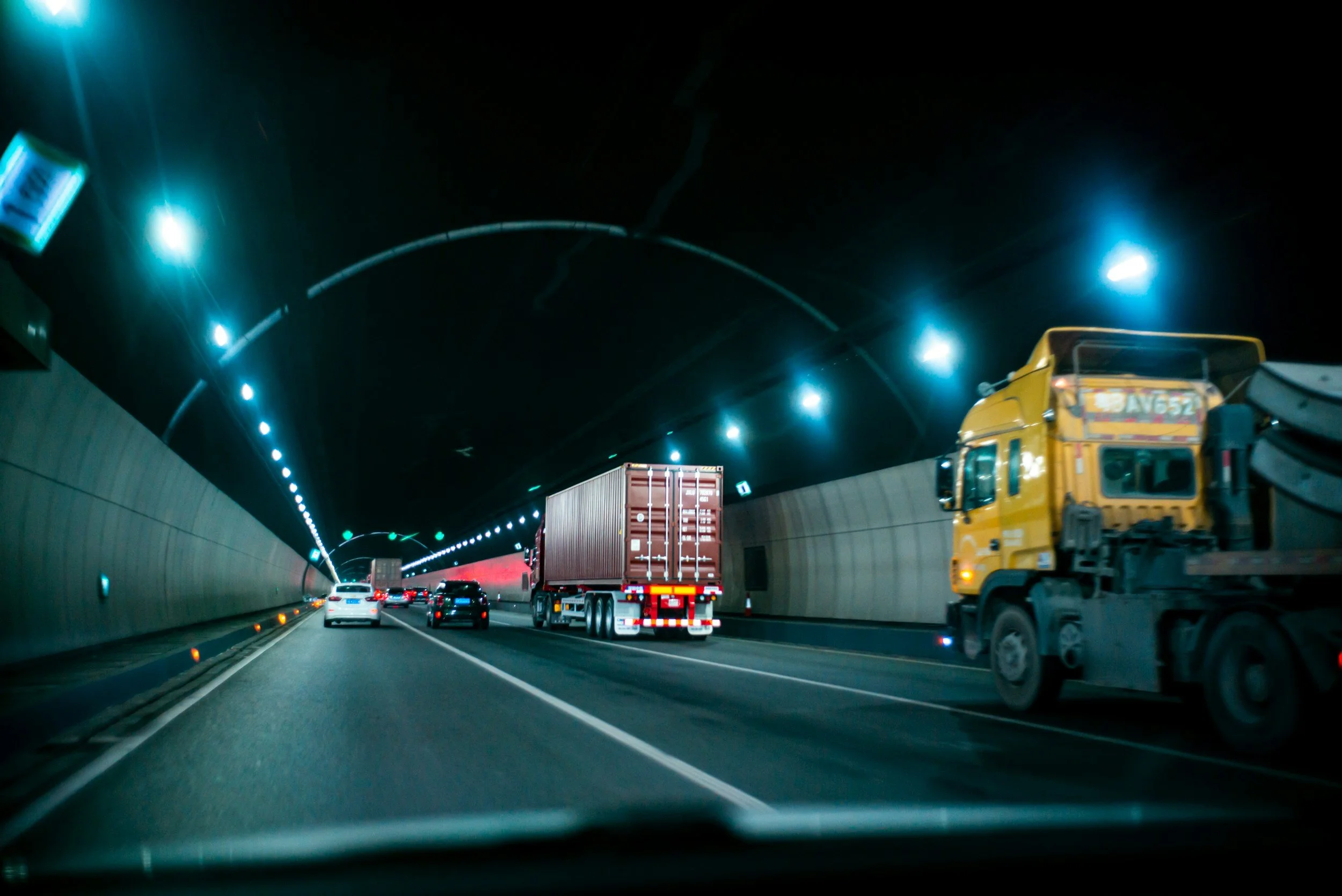 Vehicles traveling through a dark tunnel, including a yellow truck on the right, a red shipping container truck in the middle, and several cars ahead. The tunnel has bright blue lights along the ceiling.