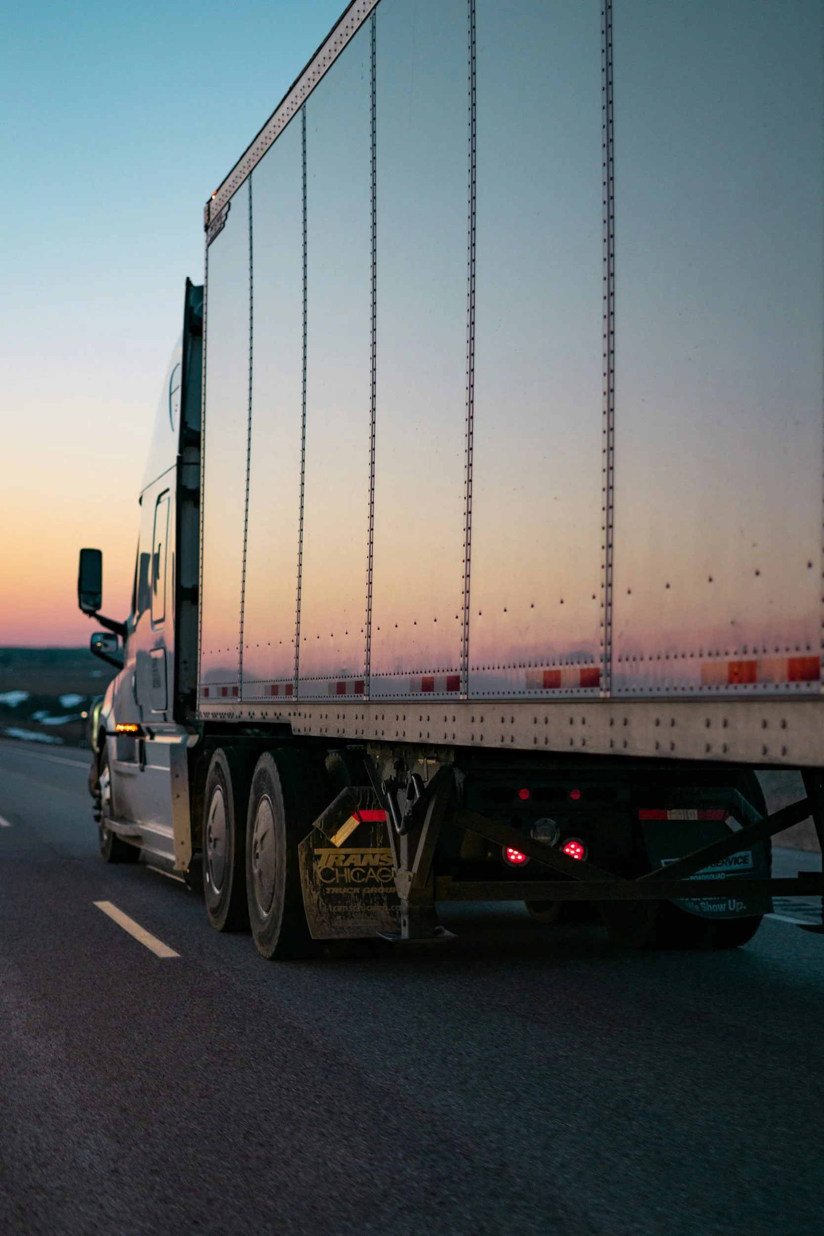 A semi-truck driving on an open highway during sunset, with a large white trailer and black wheels.