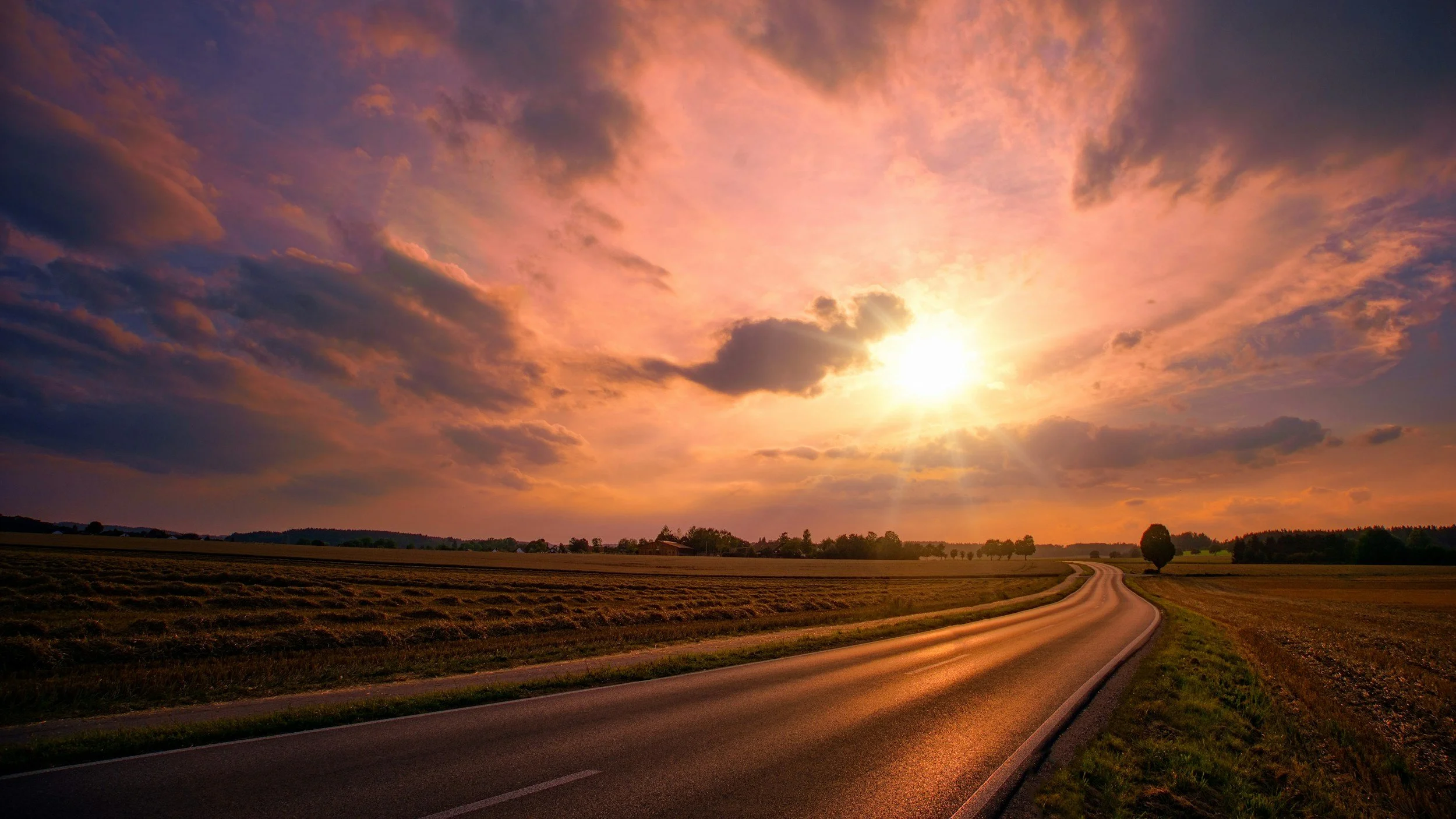 A winding country road under a colorful sunset sky with clouds and a few trees in the distance.