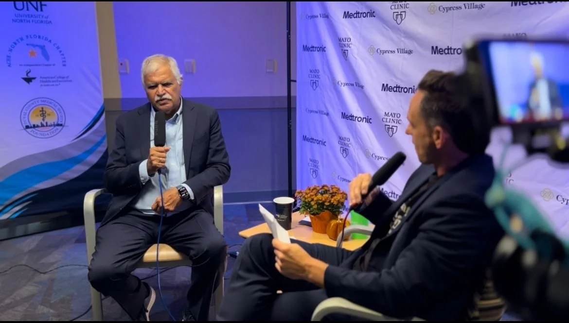 Two men in suits sitting in discussion, both holding microphones, in front of a backdrop with medical and university logos.