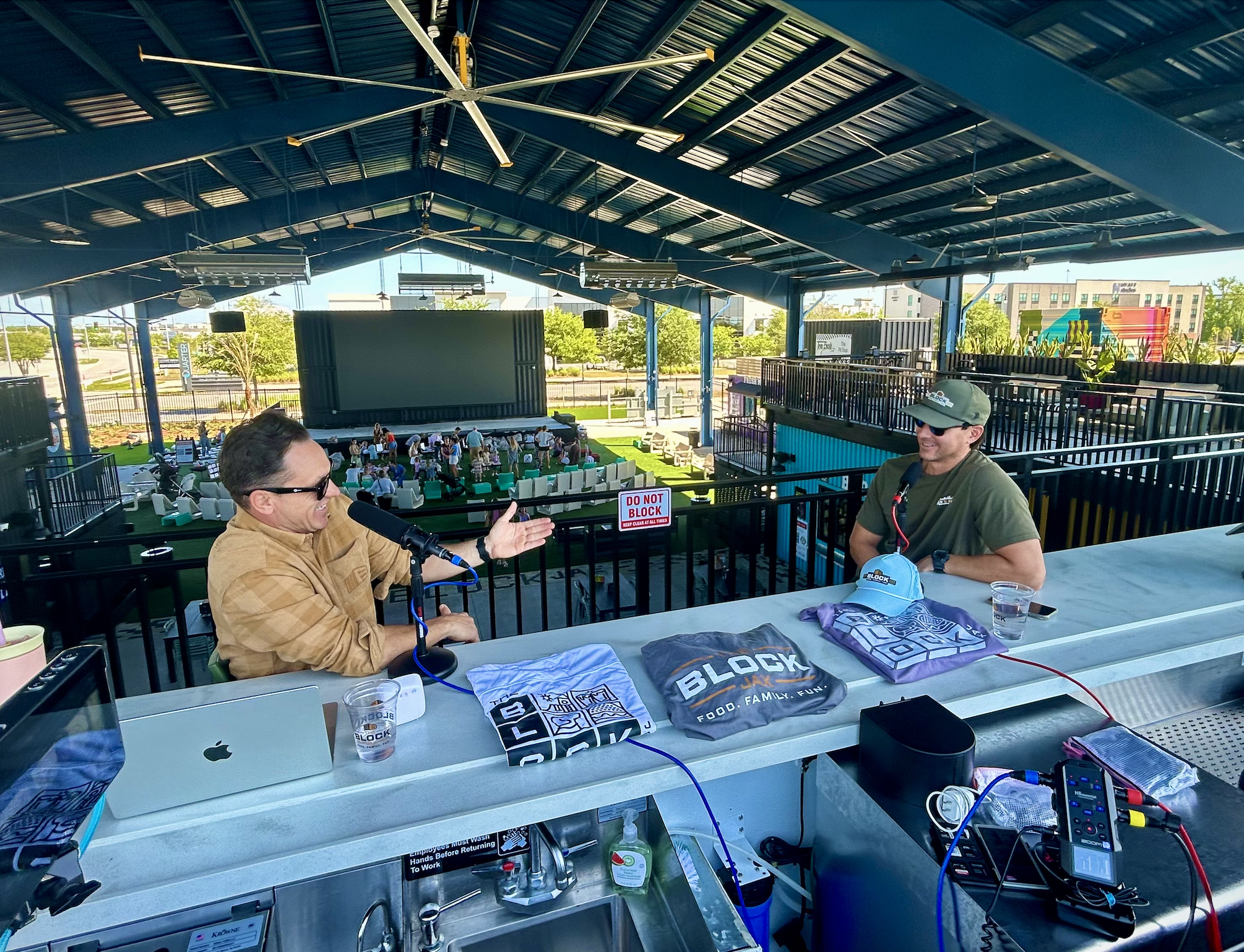 A man and woman are sitting at a table inside a blue-roofed pavilion, engaged in conversation. The table has electronic equipment, a laptop, drinks, and promotional materials. People are visible in the background, and there are signs indicating safety instructions.