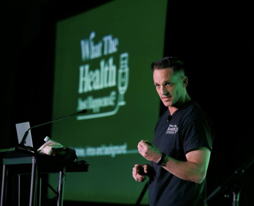 A man wearing a black T-shirt giving a presentation on stage with a large green screen behind him that reads 'We are The Health' and includes a speech bubble icon.