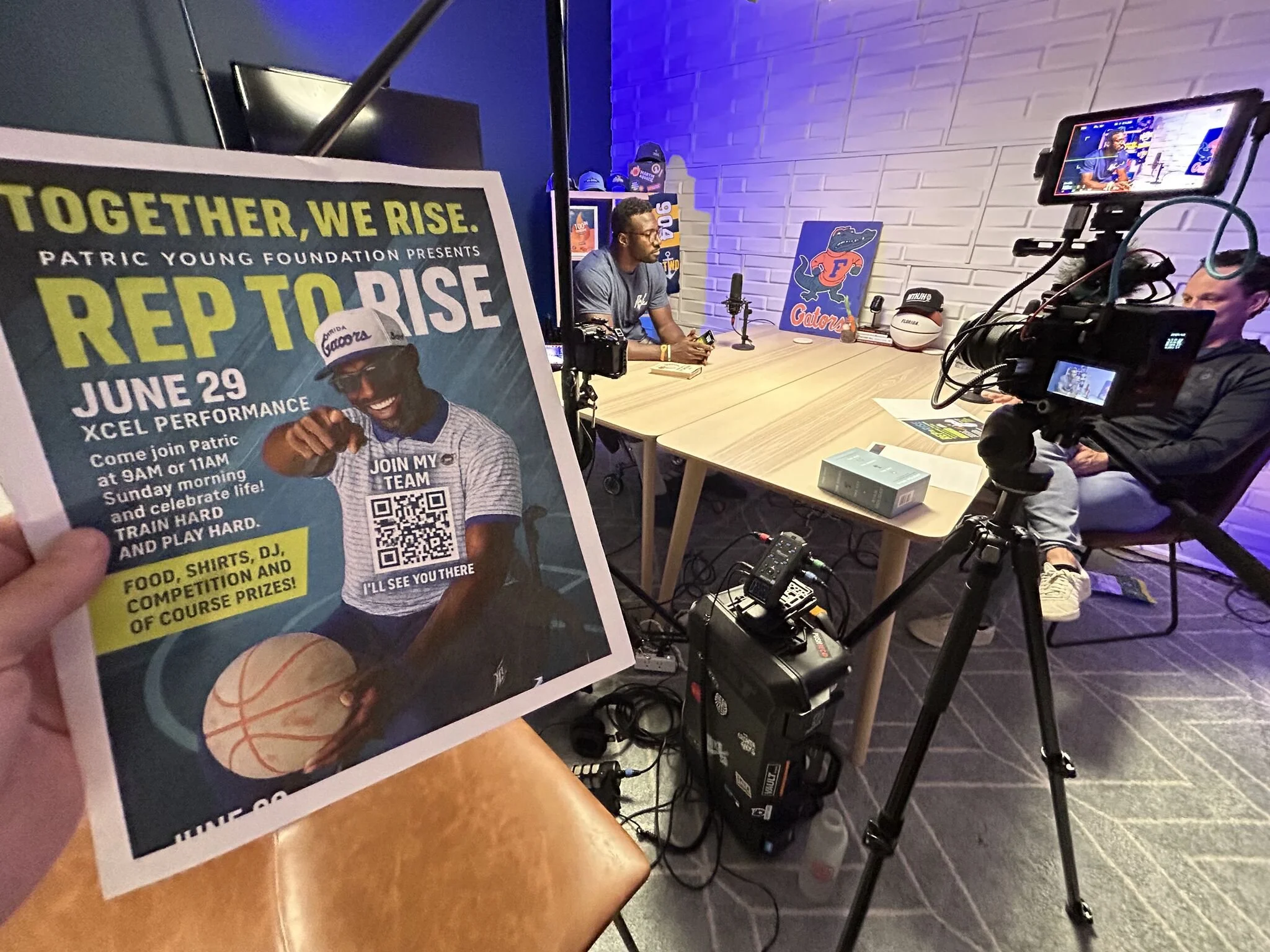 A person in a recording studio with a camera and microphone, sitting at a wooden table. In the foreground, a flyer promoting an event called 'Reboot Rise' on June 29 at the Xcel Performance is visible, featuring a smiling person and details about the event.