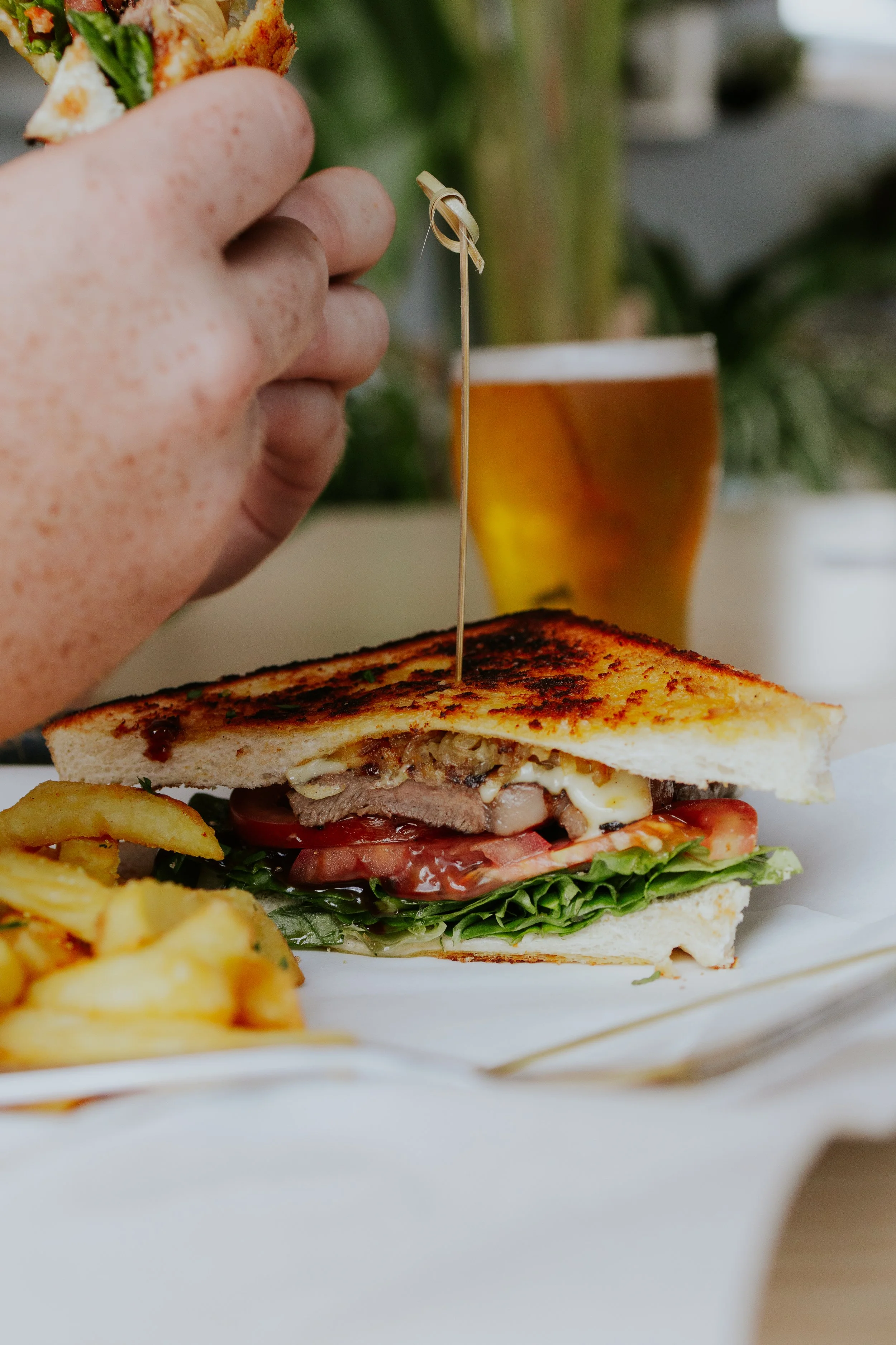 A person is holding a sandwich with grilled bread, meat, cheese, tomato, lettuce, and condiments, with a side of fries and a glass of beer in the background.