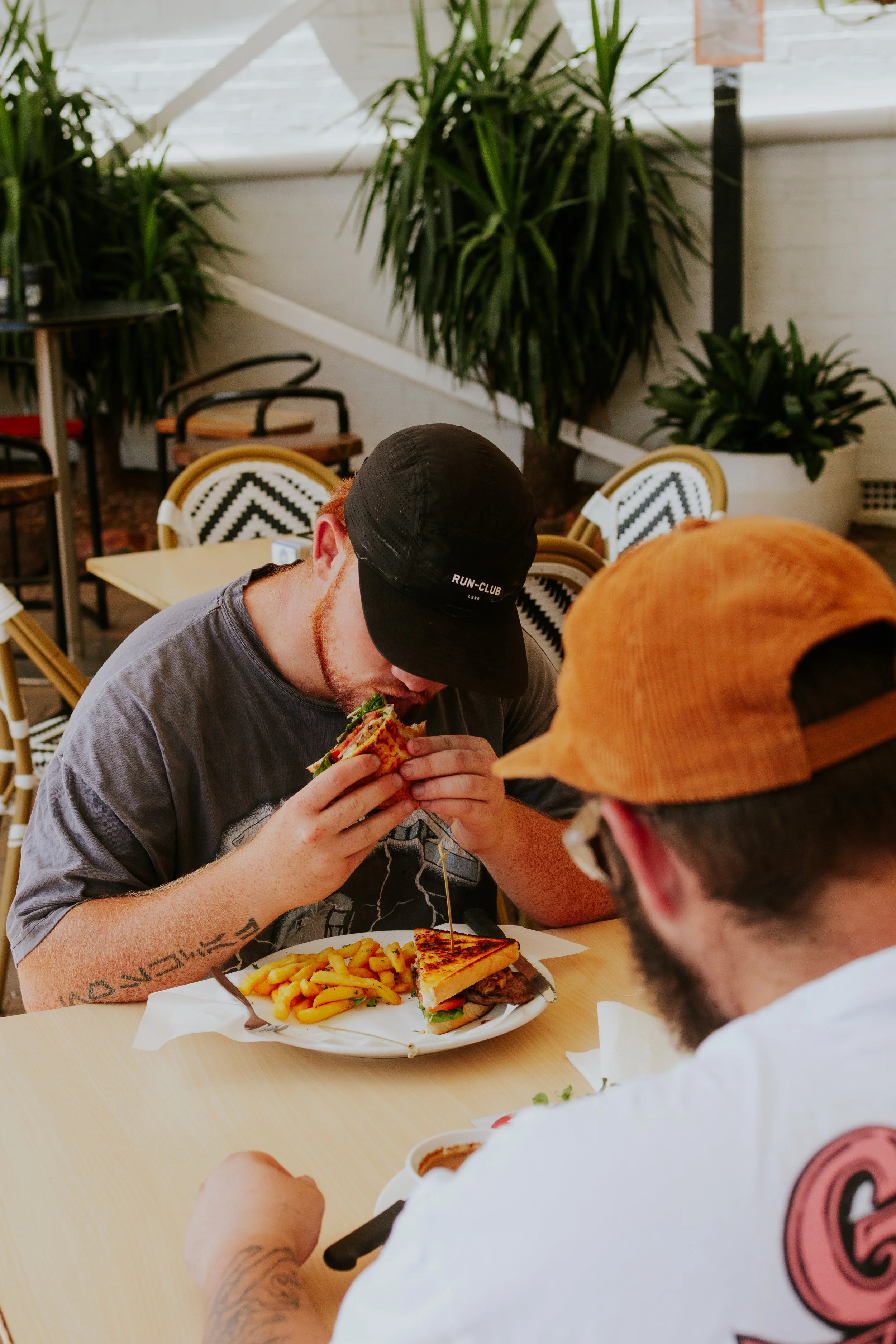 Two men seated at a restaurant table, one eating a sandwich with fries and the other with a glass of sauce, surrounded by plants and cafe chairs.