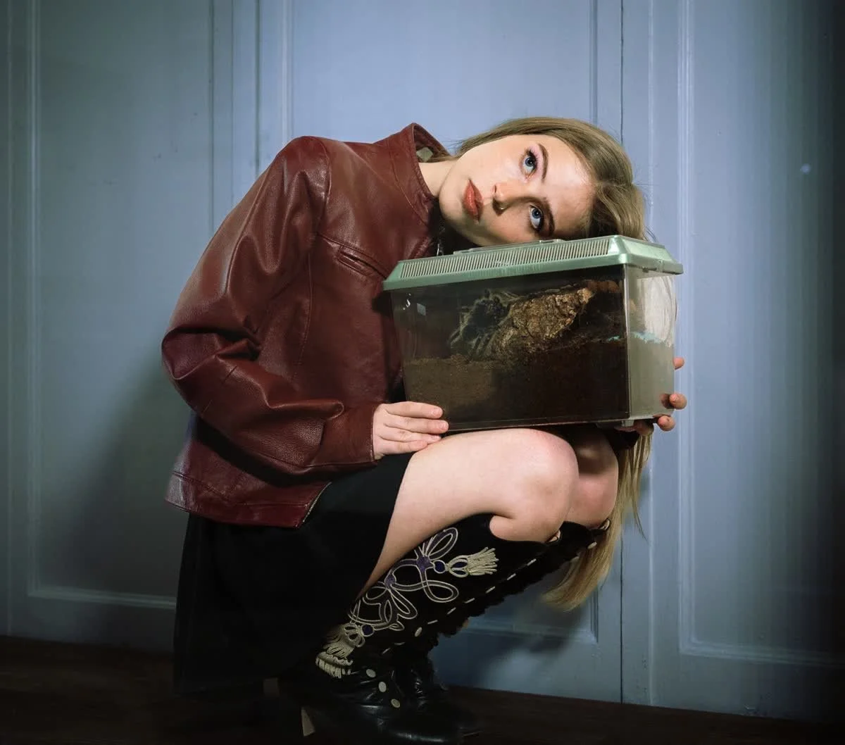 A woman with blonde hair in a dark dress and black lace-up boots crouches down in a room with blue walls, holding a terrarium with a large spider inside, and resting her head on it while looking at the camera.